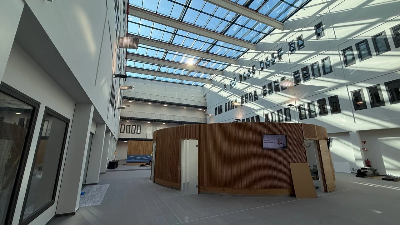 Interior of a modern building with a glass ceiling, white walls, and multiple rows of windows. There is a curved wooden structure in the center, with an open door and a small TV mounted on it. Sunlight creates shadows on the walls and floor.