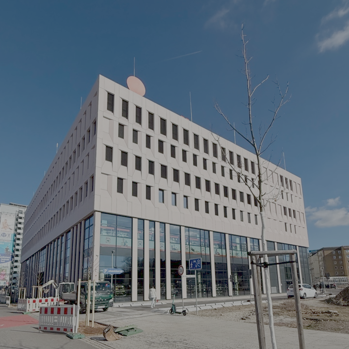 Modern multi-story building with glass windows and a unique geometric facade, with sidewalk, trees, and pedestrians in front.