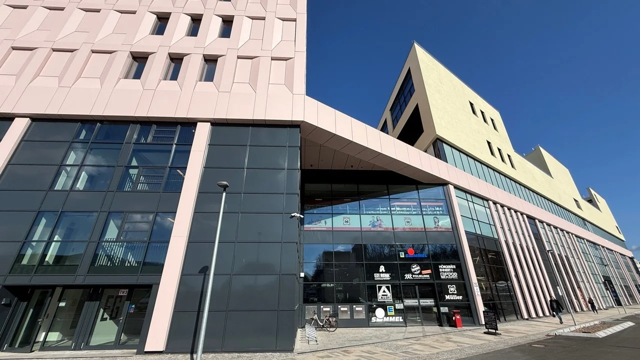 Modern shopping mall with large glass windows and various store signs, under a clear blue sky.