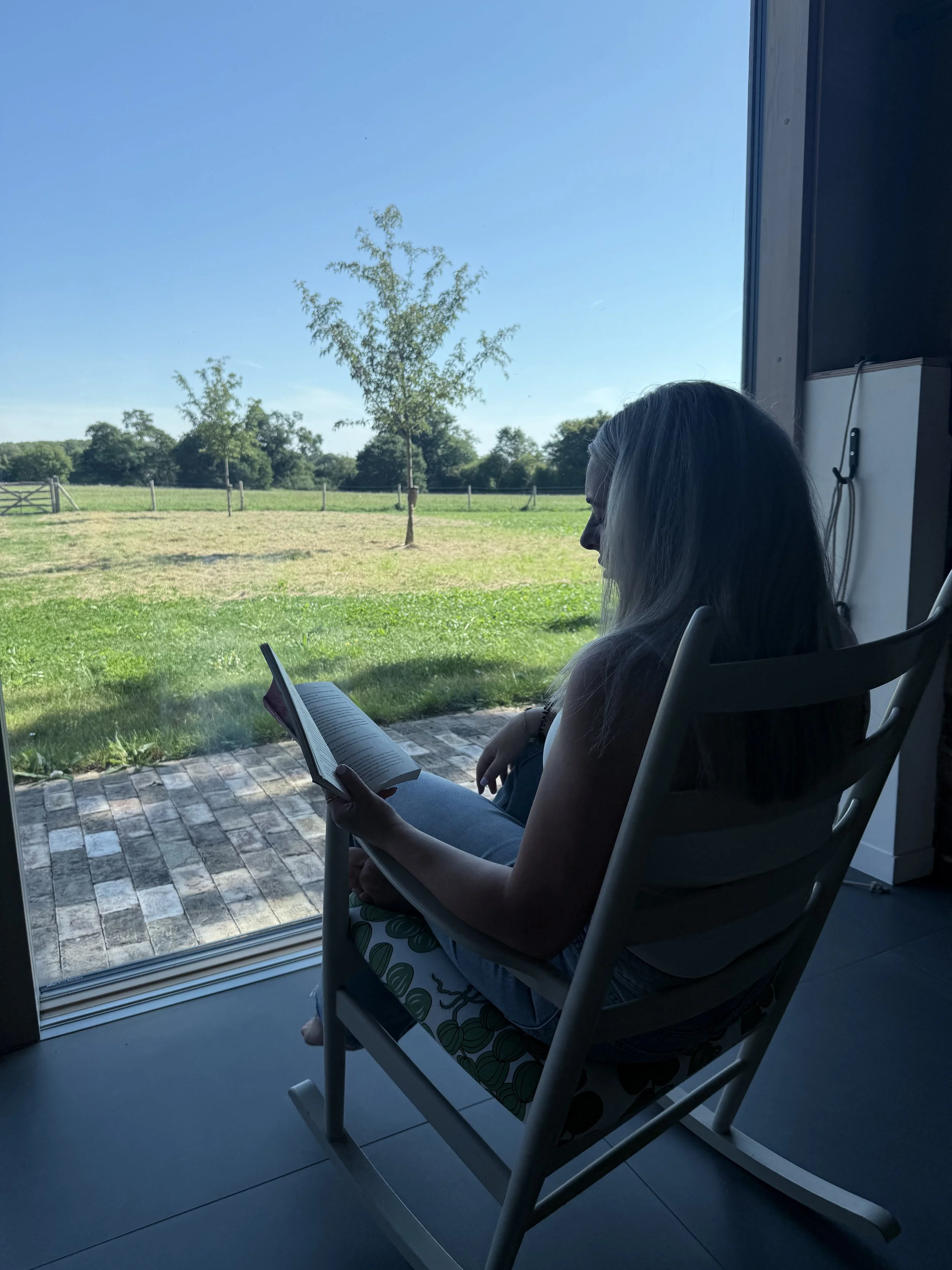 A woman with long gray hair sitting in a rocking chair inside, reading a book while looking out the window at a sunny outdoor scene with green grass, trees, and a clear blue sky.