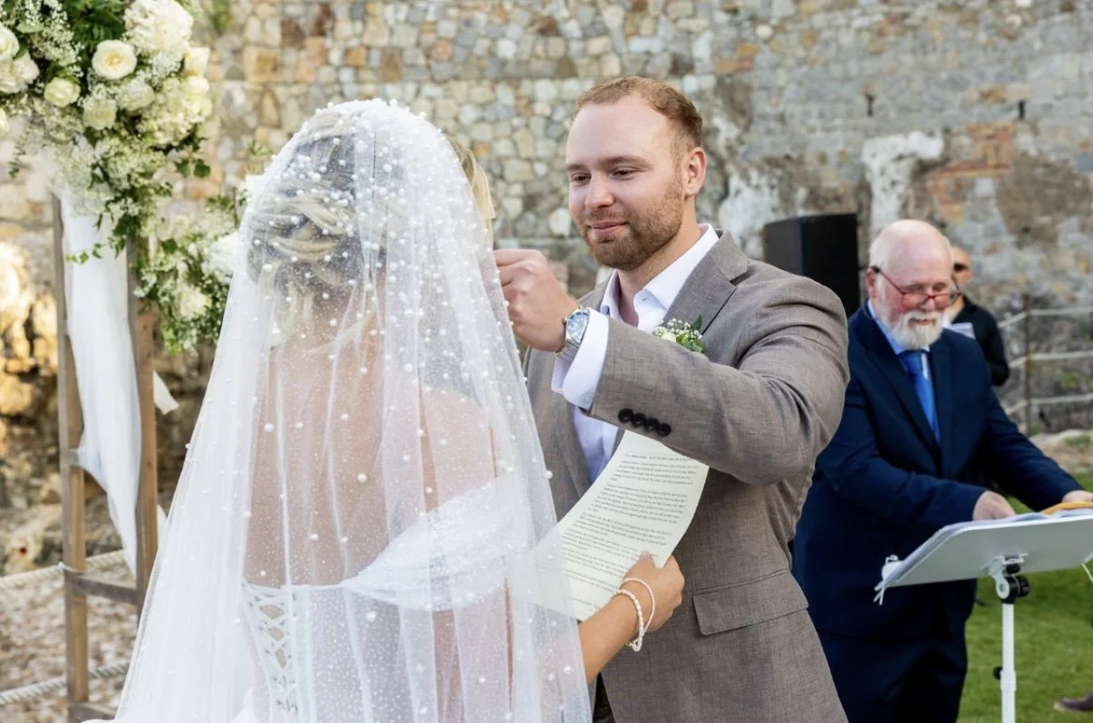 A groom is placing a ring on a bride's finger during a wedding ceremony outdoors. The bride is wearing a wedding dress and veil, while the groom is in a gray suit. Other people are present, with one older man in the background smiling.