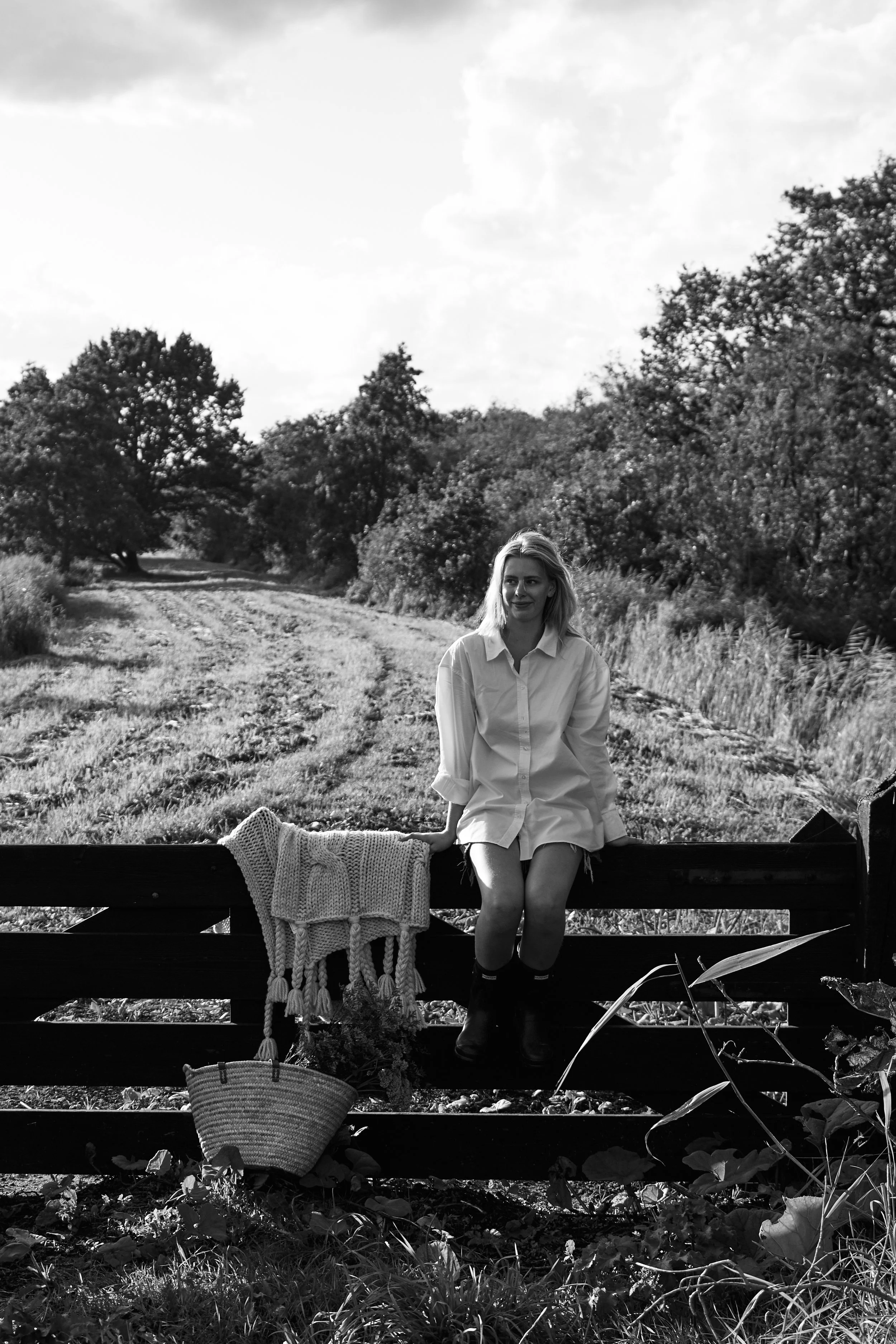 Een zwart-witfoto van een vrouw die op een houten hek zit in een landelijke omgeving met bomen en gras, met een lievelingsjas en een mand met planten voor haar. 