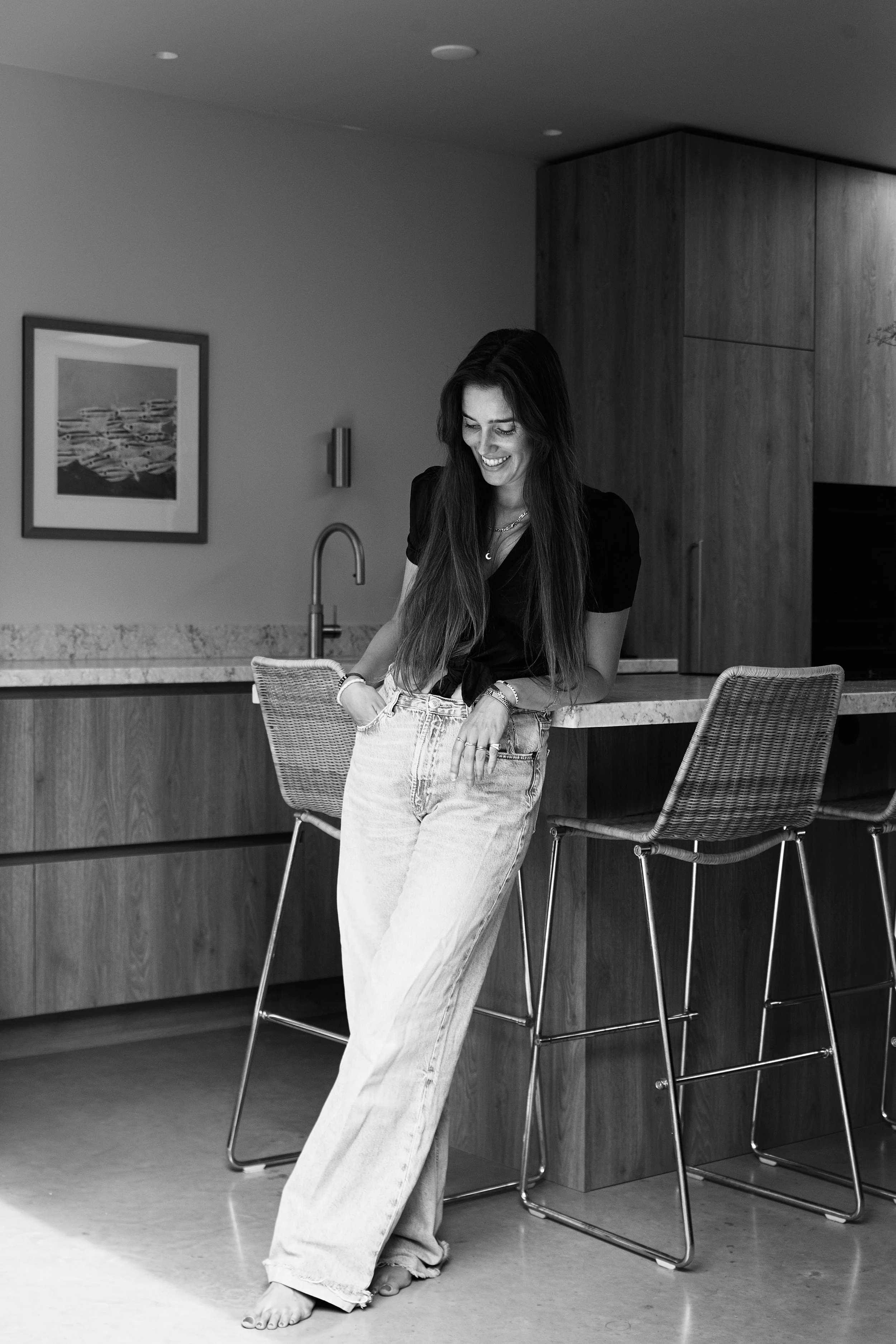 Een vrouw met lang haar die glimlacht en naar beneden kijkt, terwijl ze zonder schoenen in een modern interieur staat, met een keuken en barkrukken op de achtergrond, in zwart-wijn.