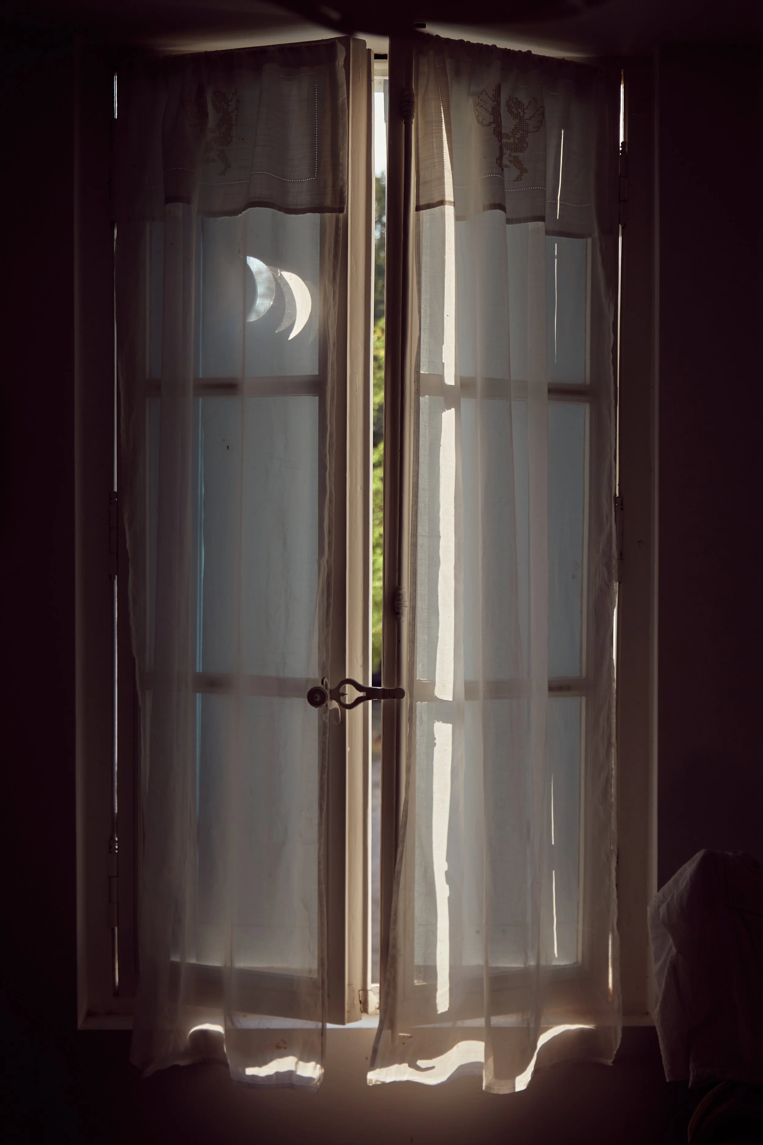 Binnenzicht van een raam met witte gordijnen, waarop door de lichte opening een maan en maansikkels zichtbaar zijn.