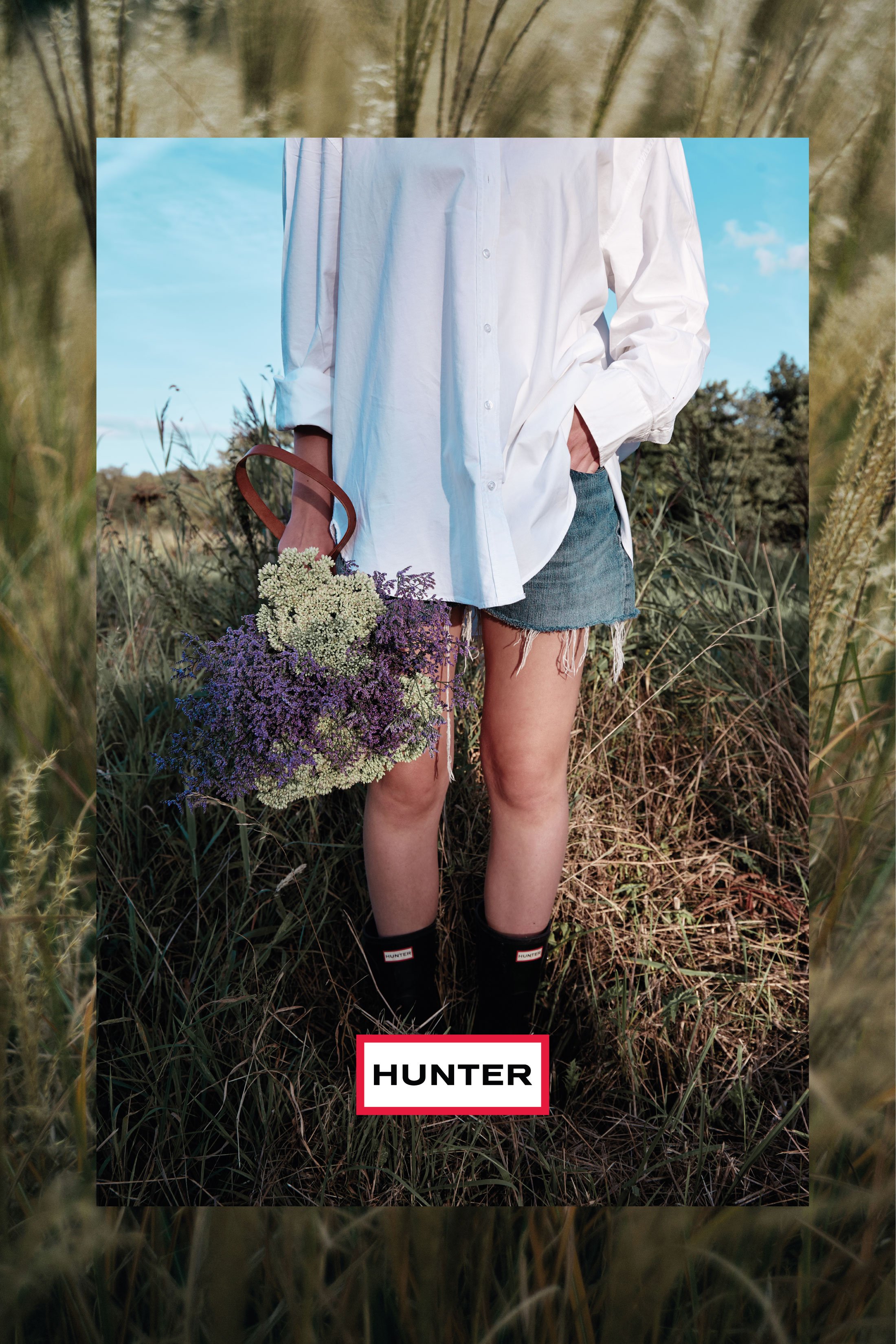 Afbeelding van een persoon die een grote bos bloemen vasthoudt, gekleed in een witte blouse met wijde mouwen, een denim rokje, en zwarte sokken met de merknaam "HUNTER". De persoon staat in een grasveld met een blauwe lucht op de achtergrond.