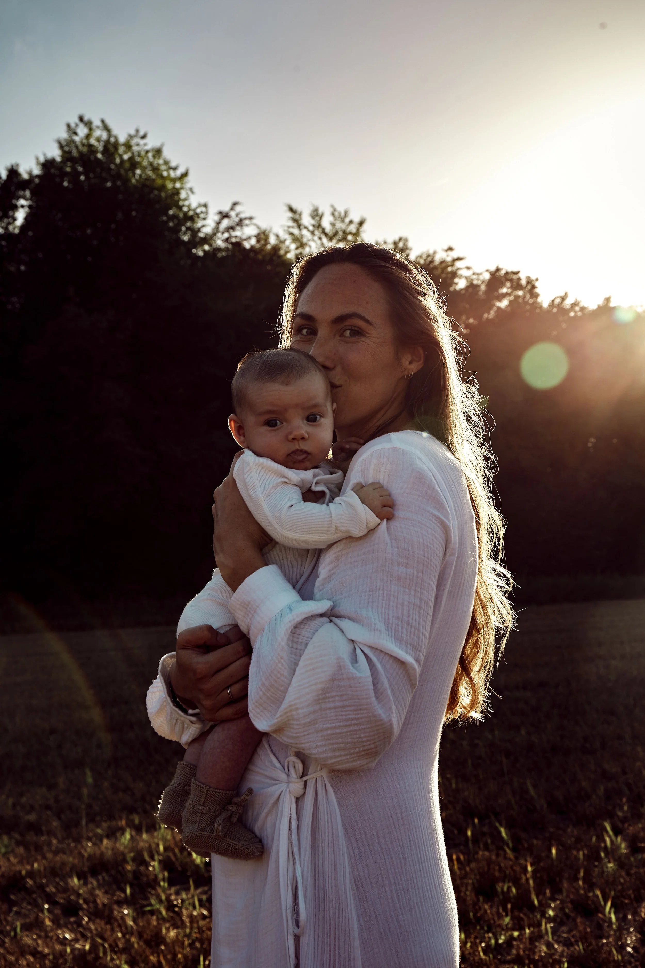 Een vrouw houdt een baby in haar armen tijdens zonsondergang in een open veld met bomen op de achtergrond.