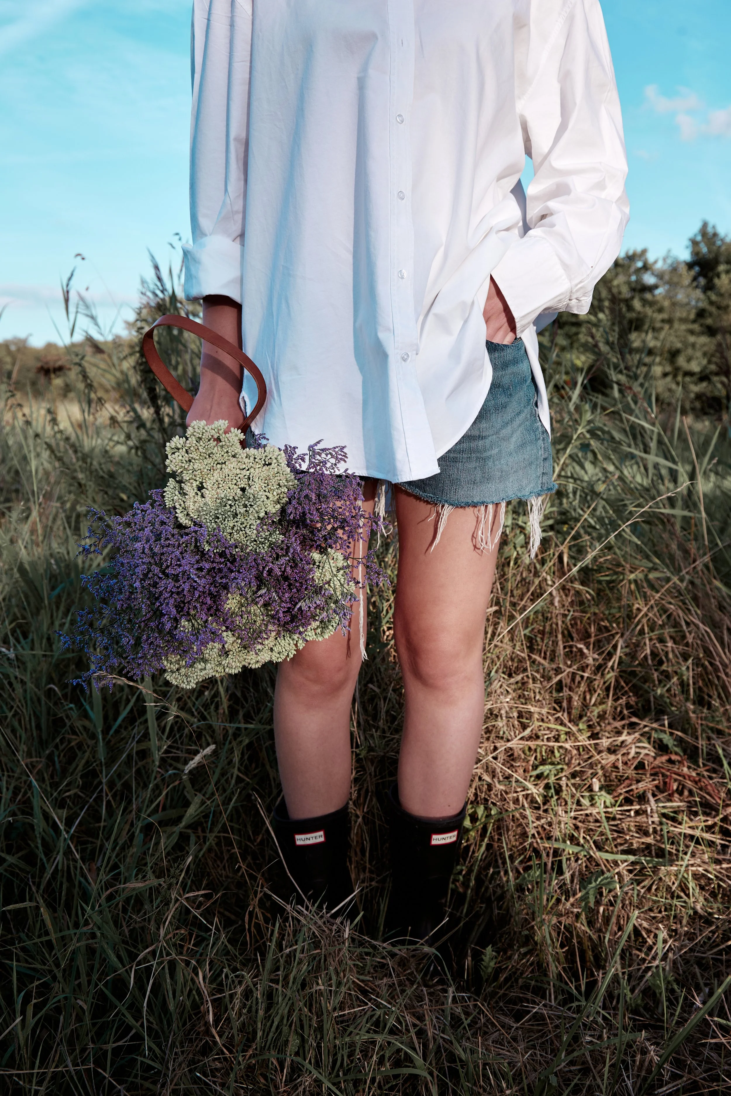 Persoon in een wit overhemd en denim shorts, die een bos bloemen vasthoudt, staat in een grasveld met een blauwe lucht op de achtergrond.