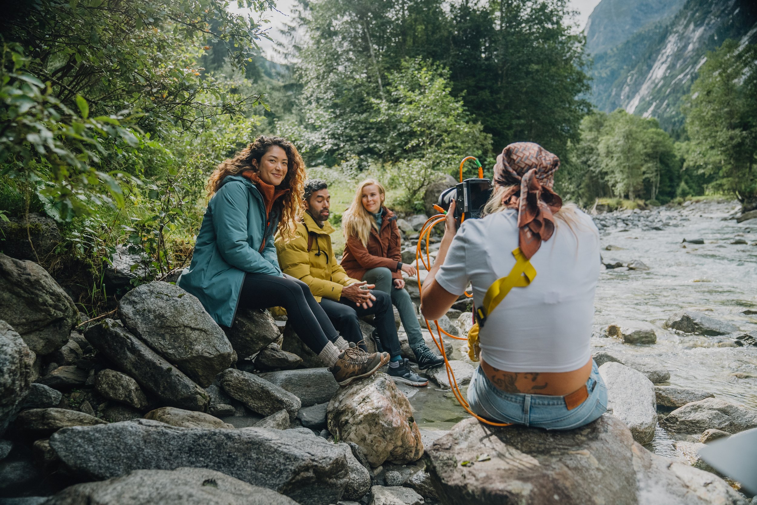 Vrouw in witte T-shirt en blauwe jeans die een fotoshoot doet van drie mensen bij een rivier in een bosrijke omgeving met bergen op de achtergrond.