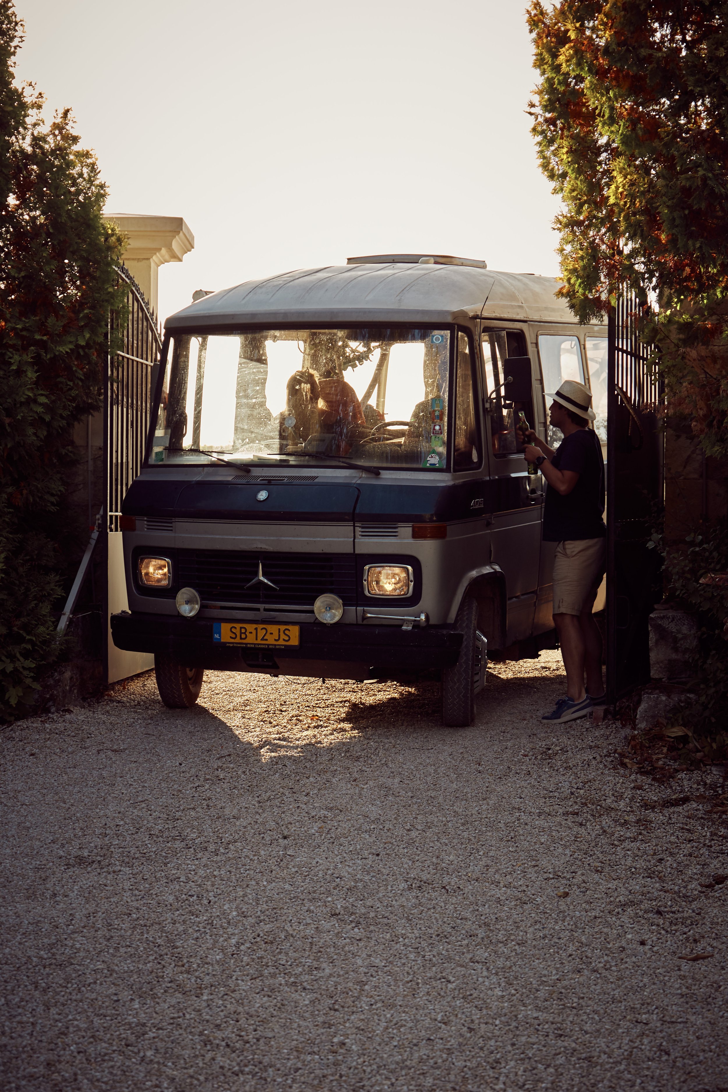 Een oude Mercedes-bus geparkeerd bij een poort, met mensen erin en erbuiten, en een vrouw die een drankje vasthoudt naast de bus bij schemerlicht.