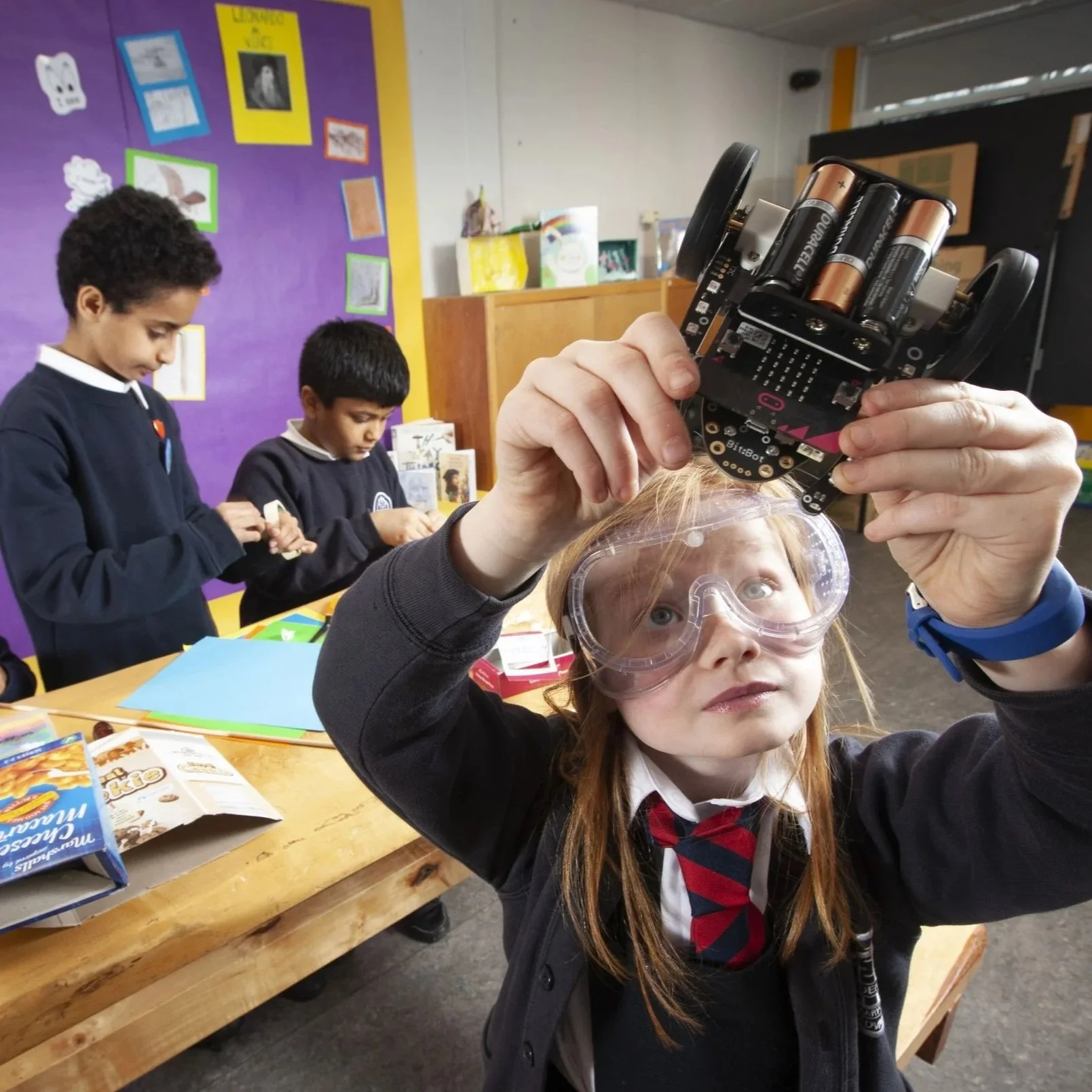 A young girl with red hair wearing safety goggles and a school uniform working with a small robot with batteries, as she looks at it closely in a classroom setting. Two boys are visible in the background, working at a table with various supplies.