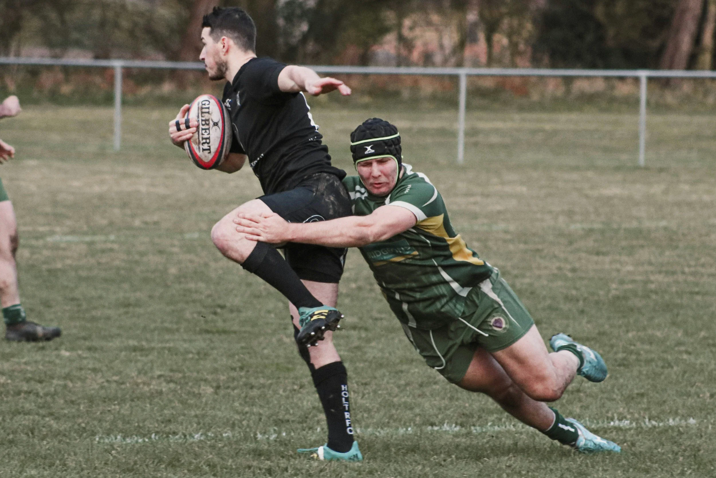 A rugby player in a black jersey holding a rugby ball runs on the field while a player in a green jersey attempts to tackle him. The game is outdoors on a grassy field.