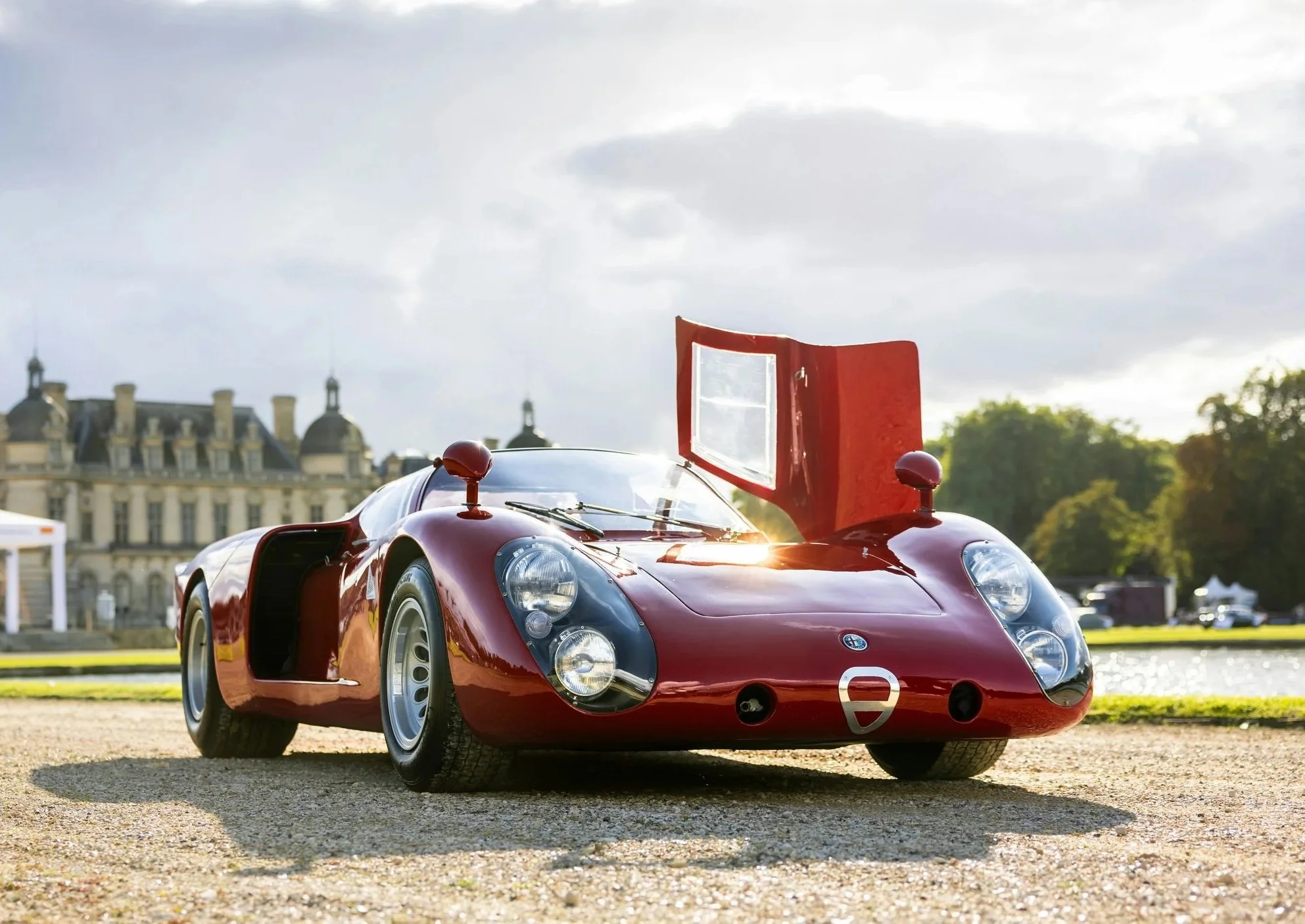 Red vintage race car with open roof and a large rear wing, parked outdoors on a gravel surface with historic buildings and trees in the background.
