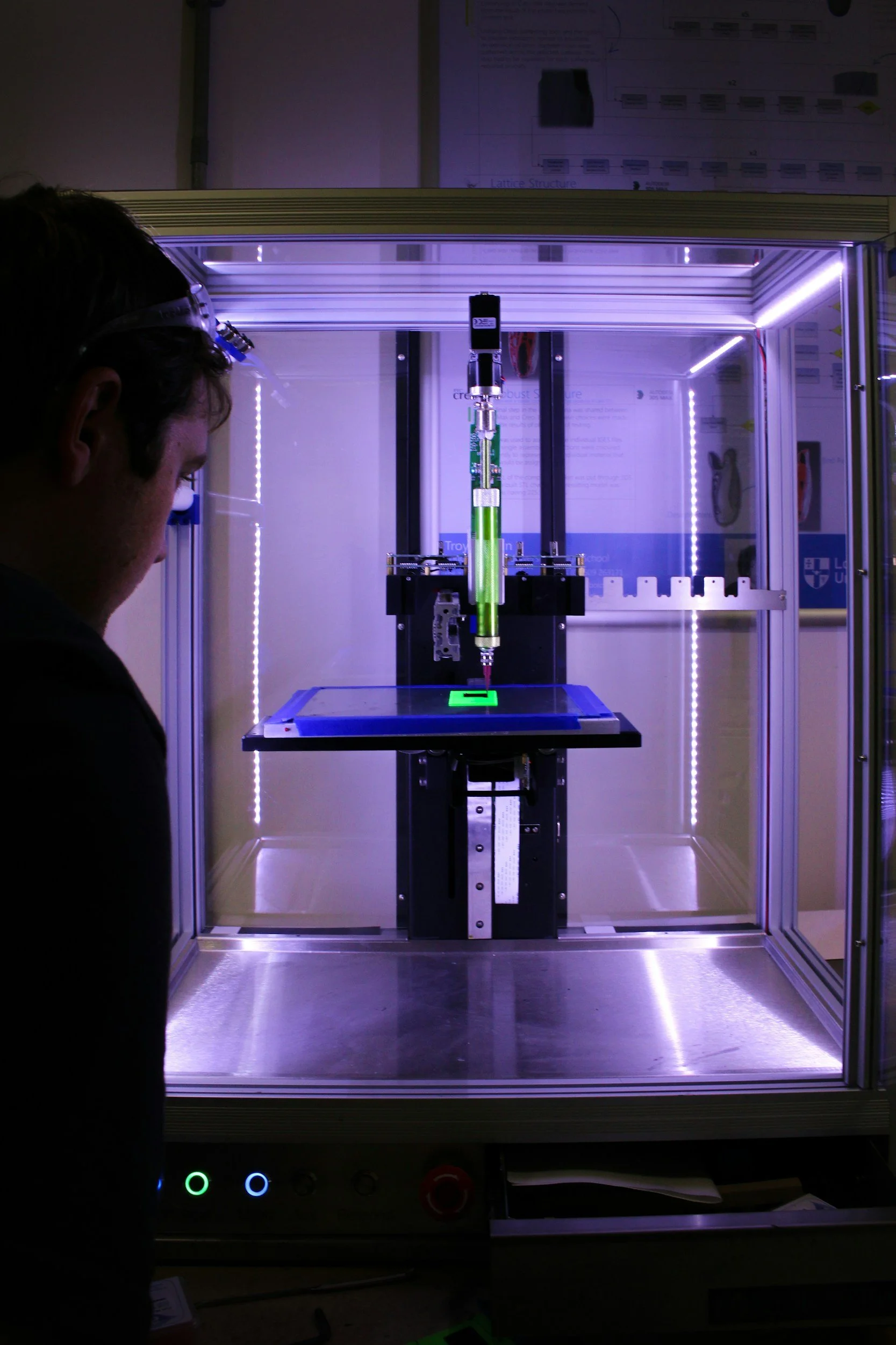A person wearing safety goggles watches a robotic device with a green-lit laser engraver inside a transparent enclosure, working on a small green object on a black platform.