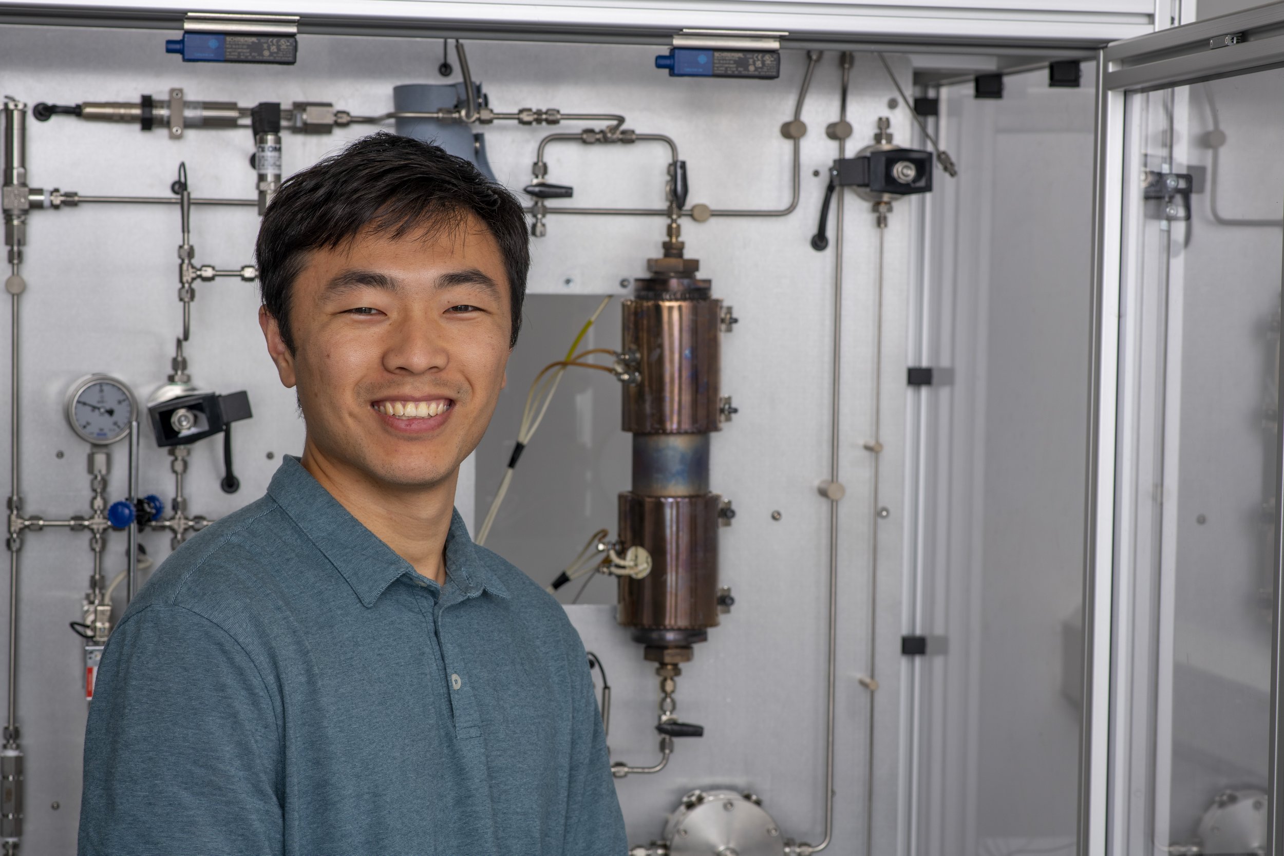 A smiling man in a blue shirt standing in front of scientific laboratory equipment with pipes, gauges, and a copper coil.