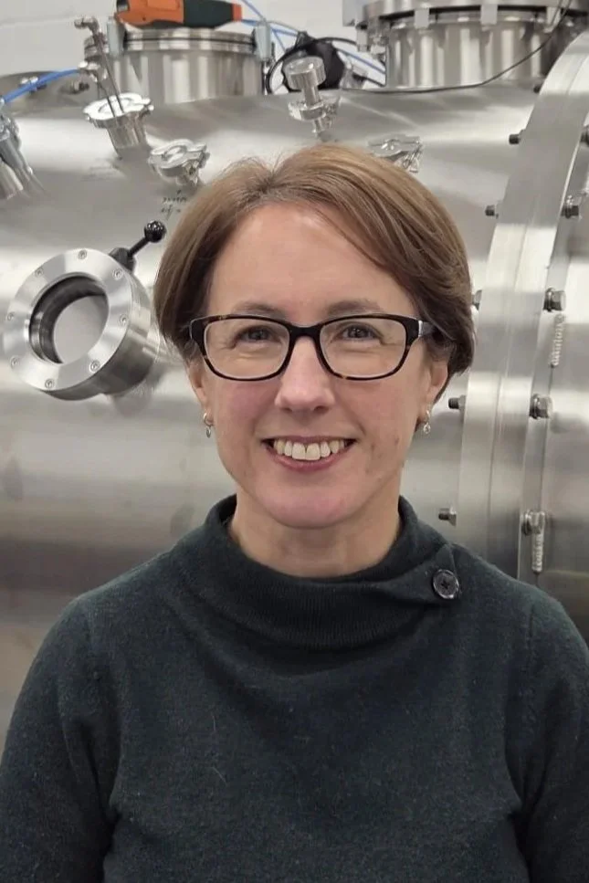 A woman with short brown hair and glasses smiling in front of a large metallic scientific instrument or machinery.