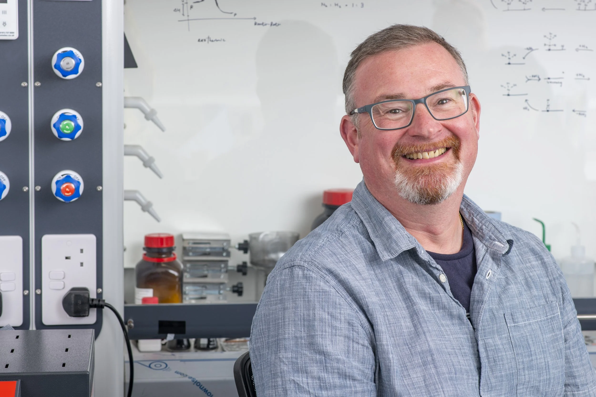 A smiling man with glasses and a beard in a laboratory setting with scientific equipment and a whiteboard with diagrams in the background.