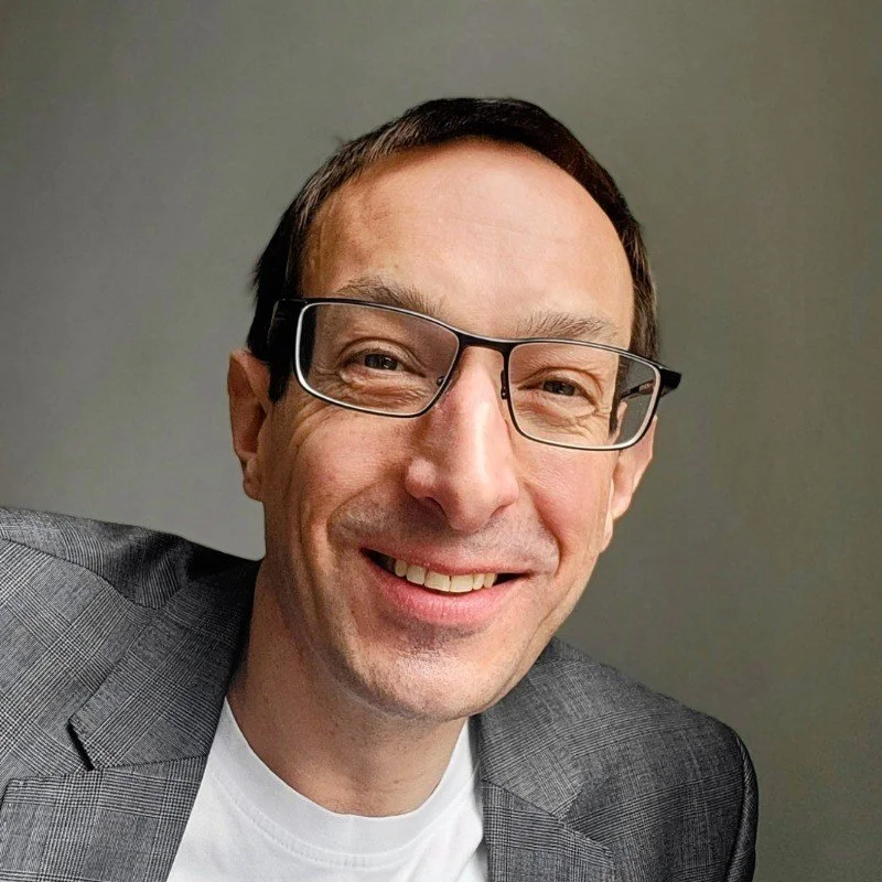 Close-up of a smiling man wearing glasses, a gray blazer, and a white T-shirt against a plain background.