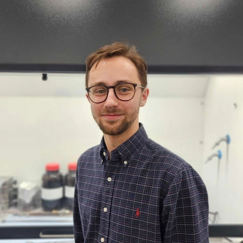 A young man with light skin, glasses, short brown hair, and a beard, smiling, wearing a dark checkered collared shirt, in a laboratory setting with shelves and bottles in the background.