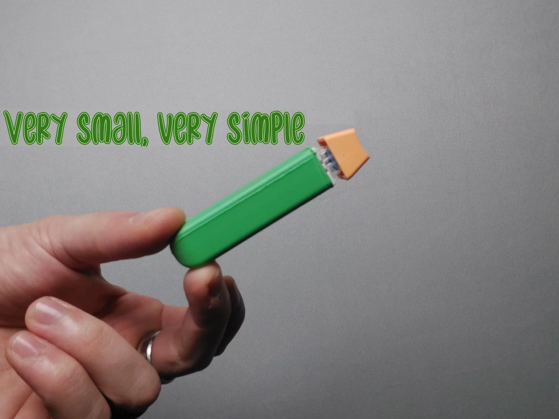 A hand holding a small green USB flash drive shaped fidget toy with a beige cap, positioned against a plain gray background. Green text on the image reads "Very small, very simple."