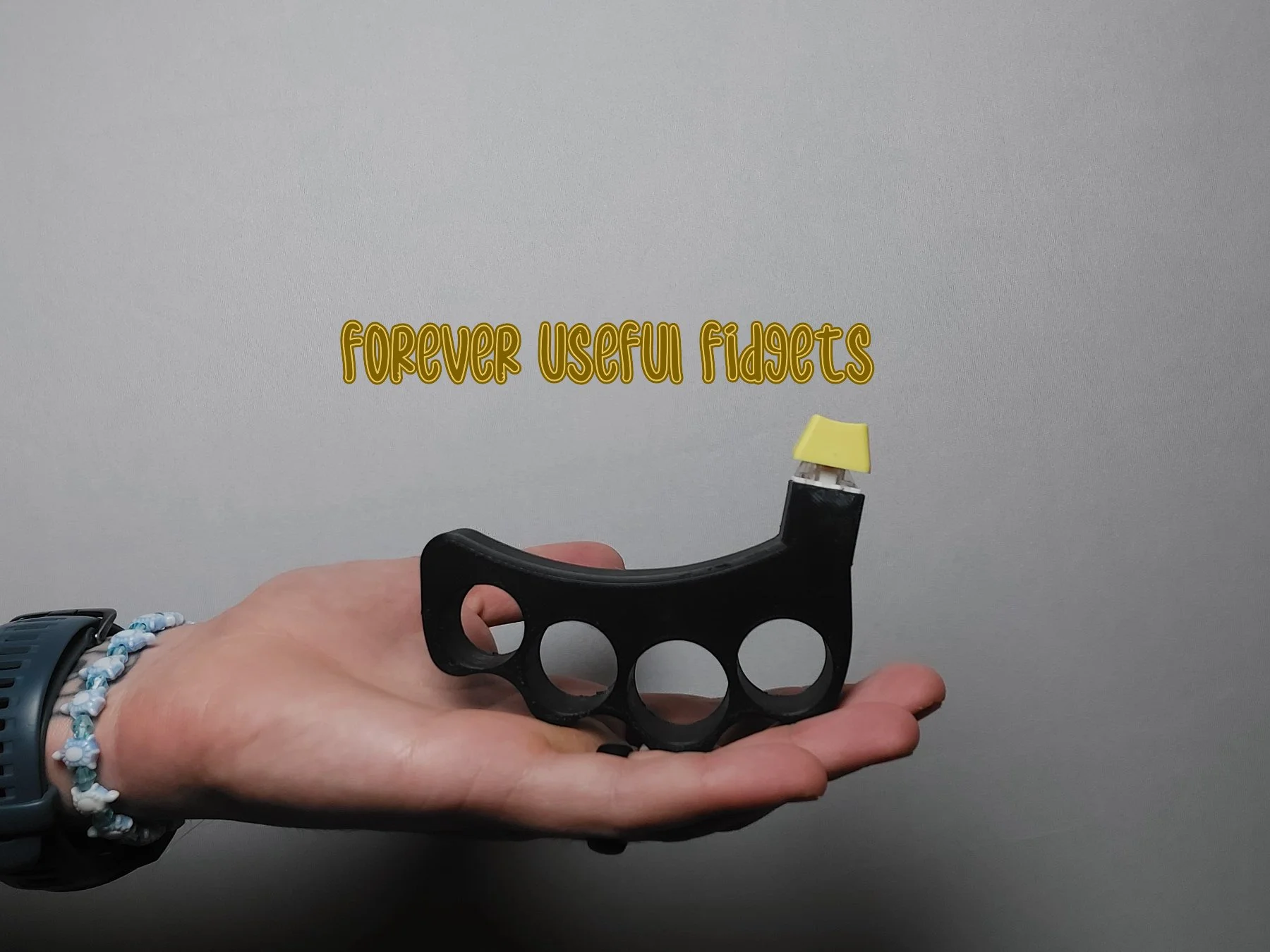 A hand holding a brass knuckle-shaped fidget toy with a yellow piece on top and a yellow text overlay reading 'Forever Useful Fidgets'.