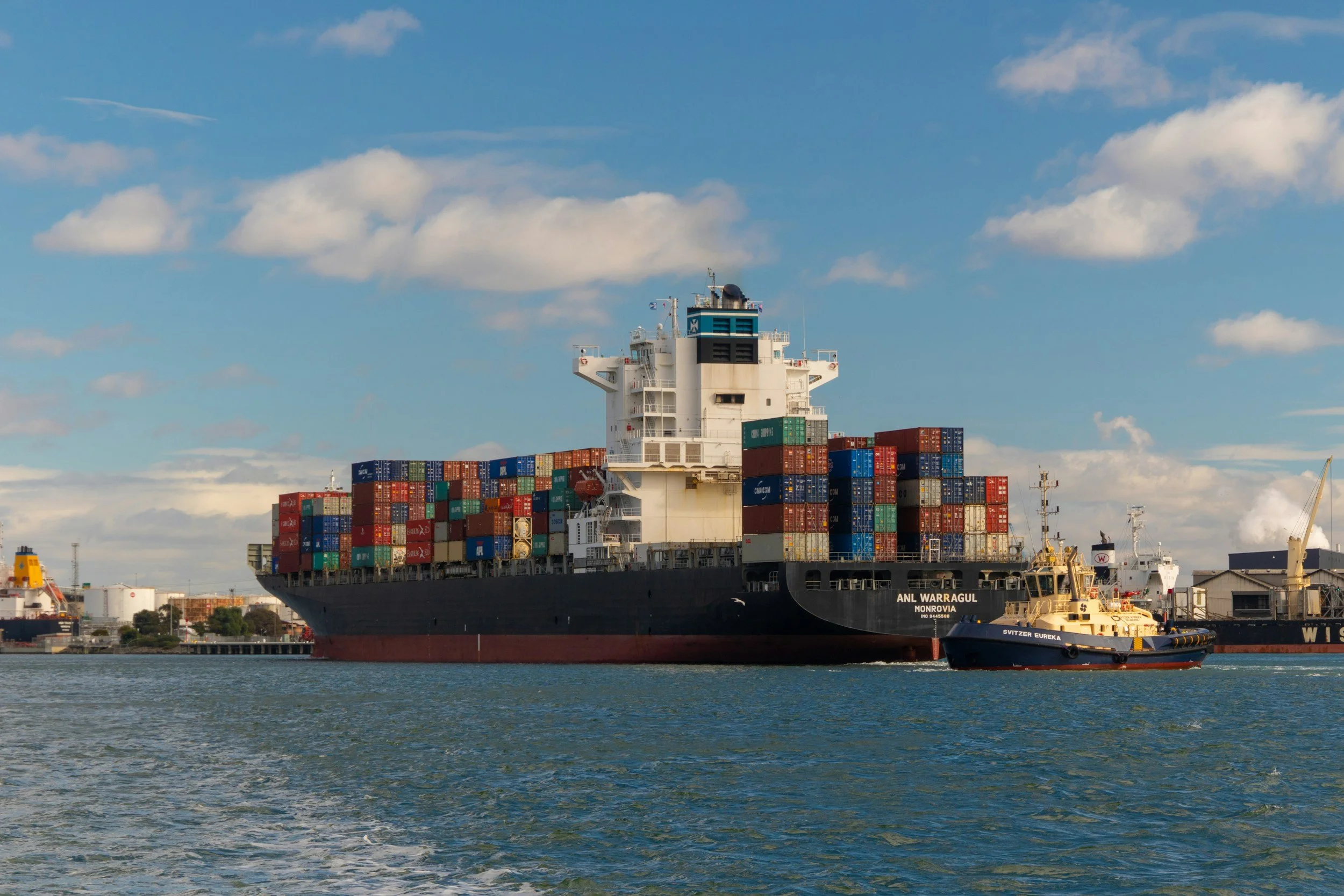 Large cargo ship with numerous stacked shipping containers sailing in a harbor, accompanied by a smaller tugboat, under a partly cloudy sky.