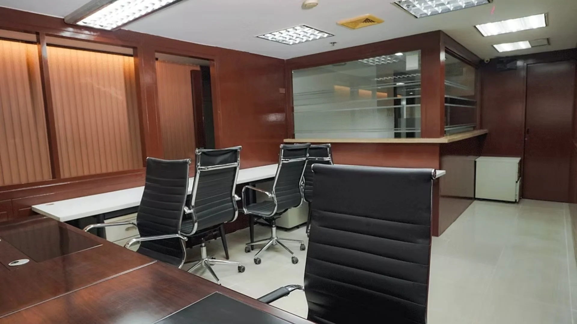 Empty office conference room with a dark wooden table, black leather chairs, wood-paneled walls, and a reception window in the background.