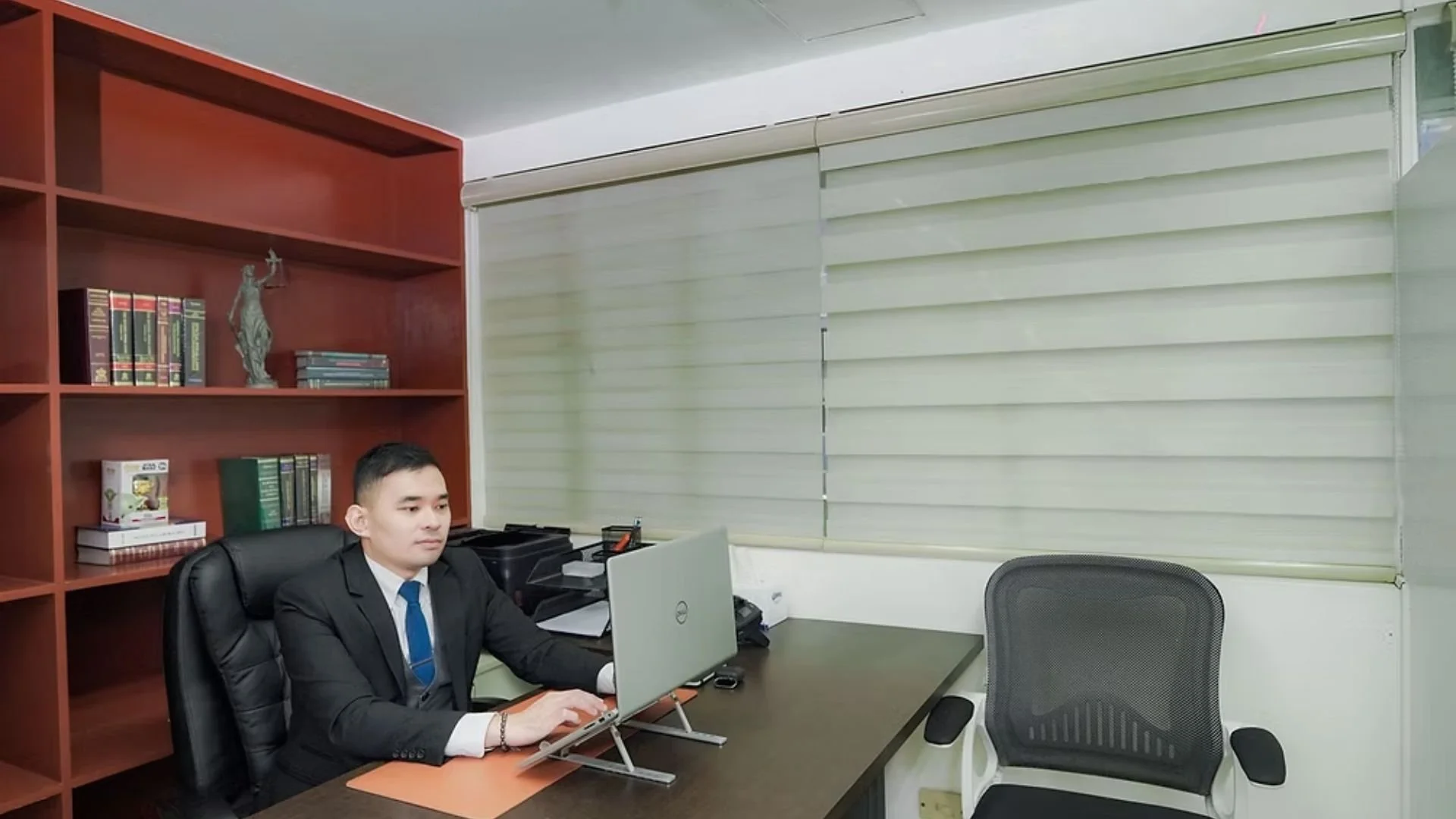 A man in a black suit and blue tie working on a computer at a desk in an office with a wooden bookshelf filled with books and a bronze statue of Lady Justice behind him. There is a window with closed blinds and an additional black office chair next t
