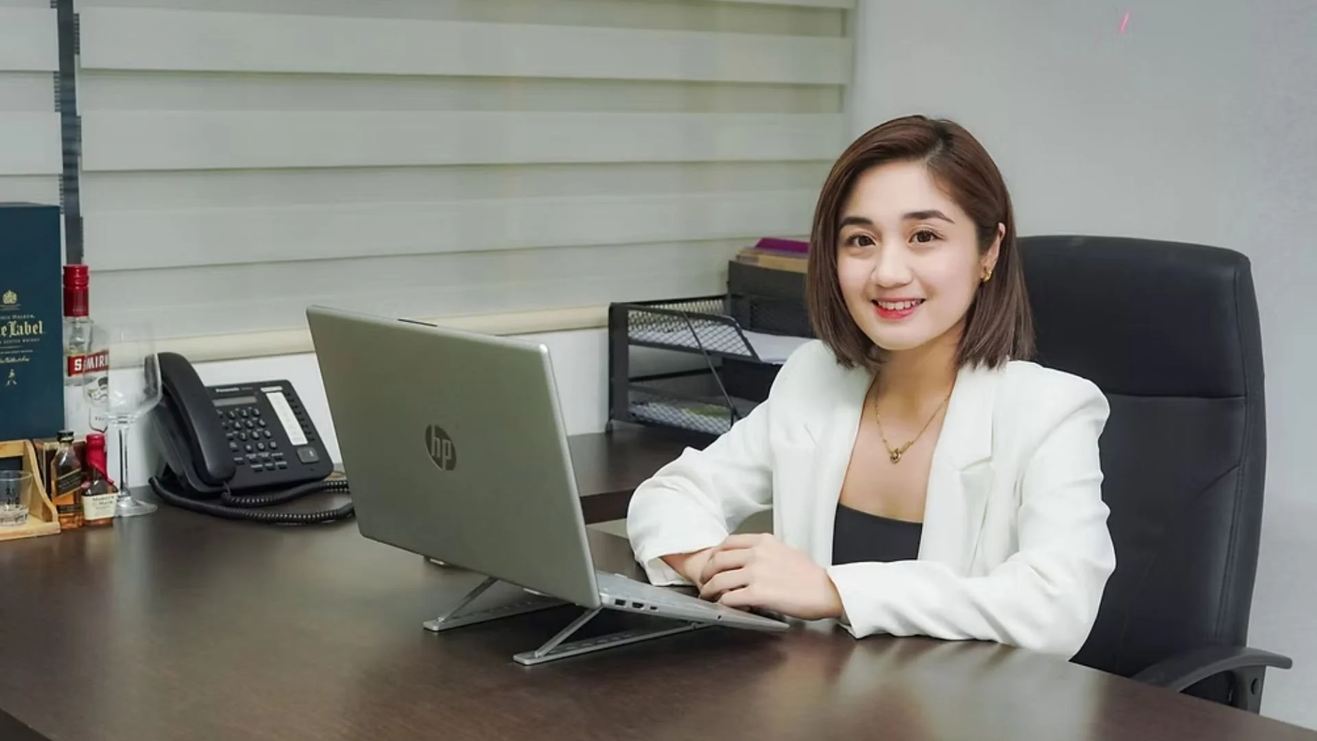 Professional woman smiling at the camera, sitting at a desk with a laptop, office phone, and files, in a modern office environment.