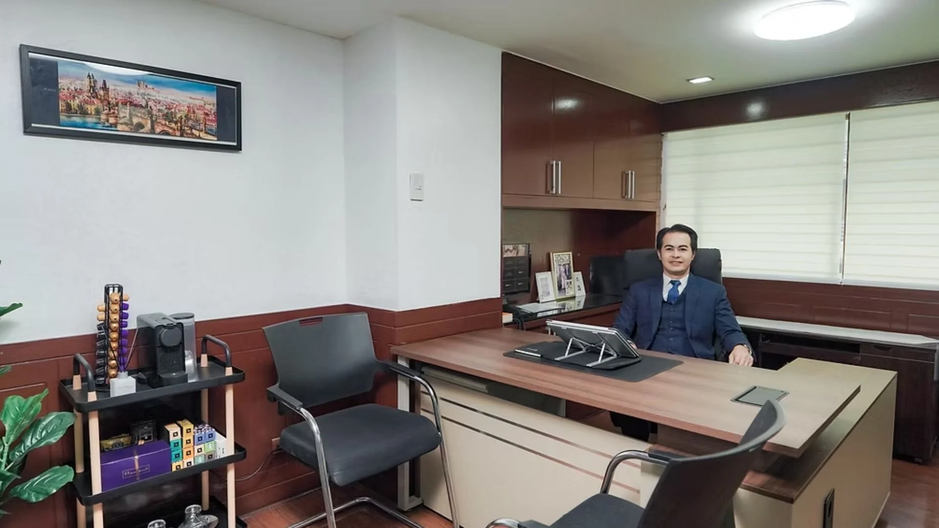 An office with a man in a blue suit sitting at a large wooden desk. The office has wooden cabinets, framed photos, a computer, and window blinds.