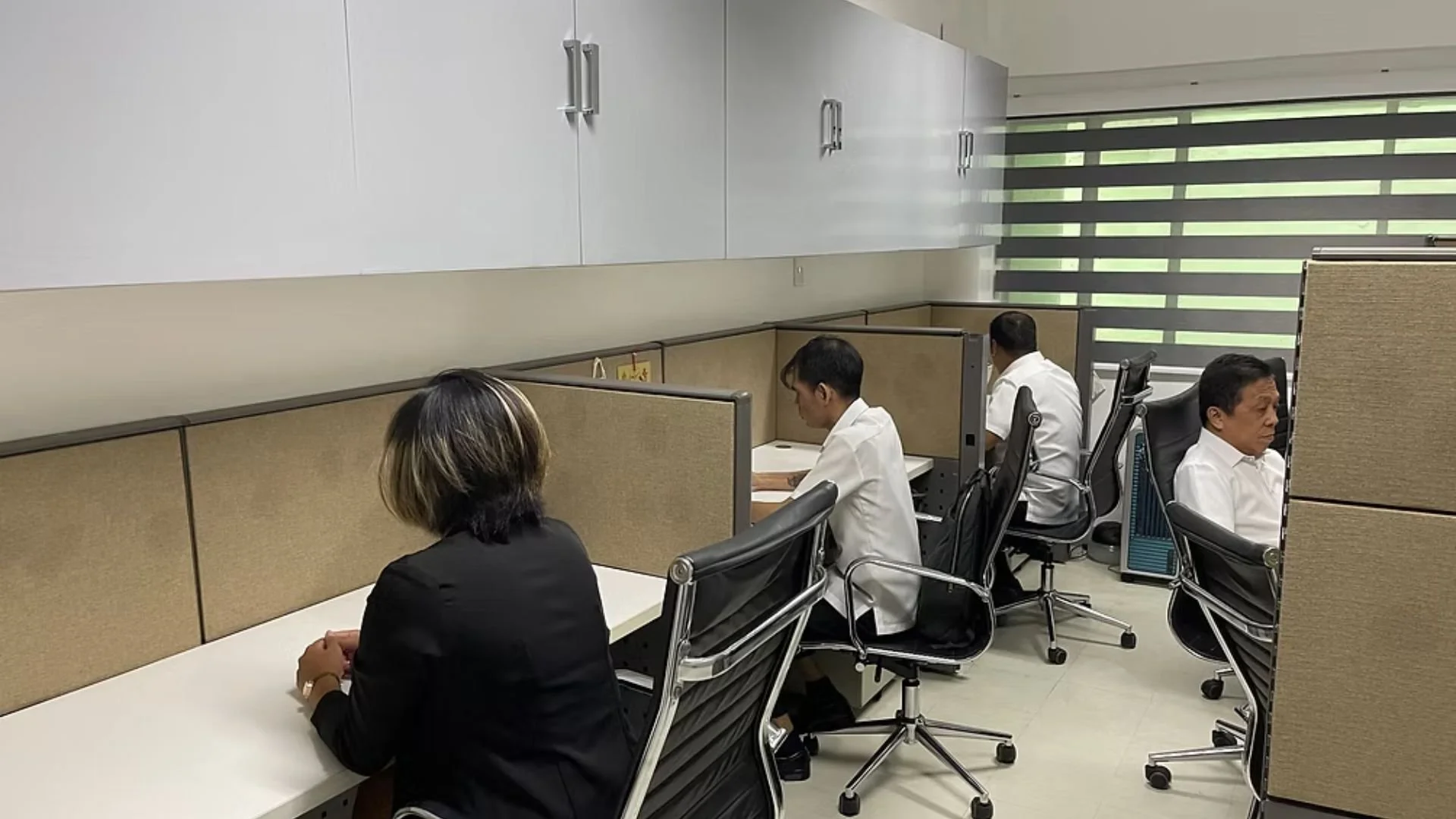 Office workers sit at their desks in cubicles, focused on their tasks in a modern office space with gray chairs and window blinds.