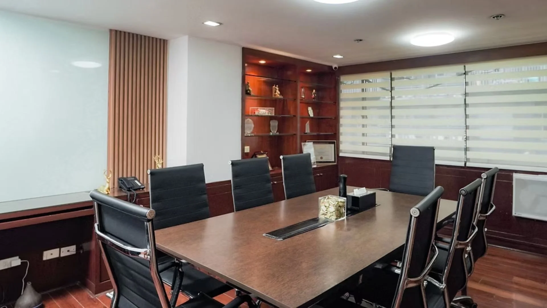 Empty conference room with a large wooden table, black chairs, built-in wooden shelves with decorative items, and windows with striped blinds.