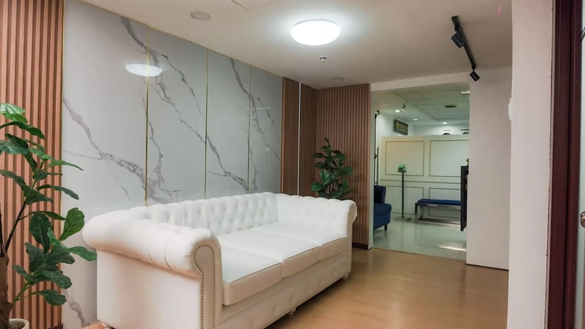 A waiting area with a white tufted sofa, potted plant, wall with marble-like panels, and a doorway leading to another room with chairs and a small table.