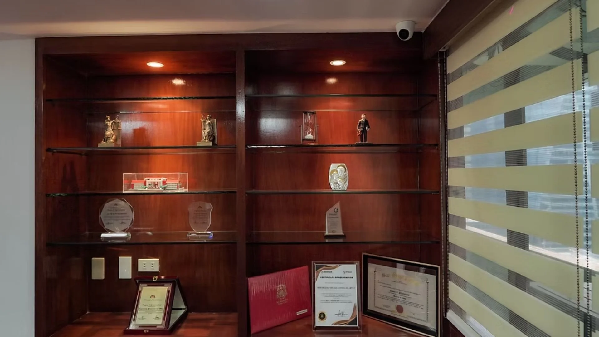A wooden display cabinet with awards, trophies, and certificates inside, next to a window with horizontal blinds.