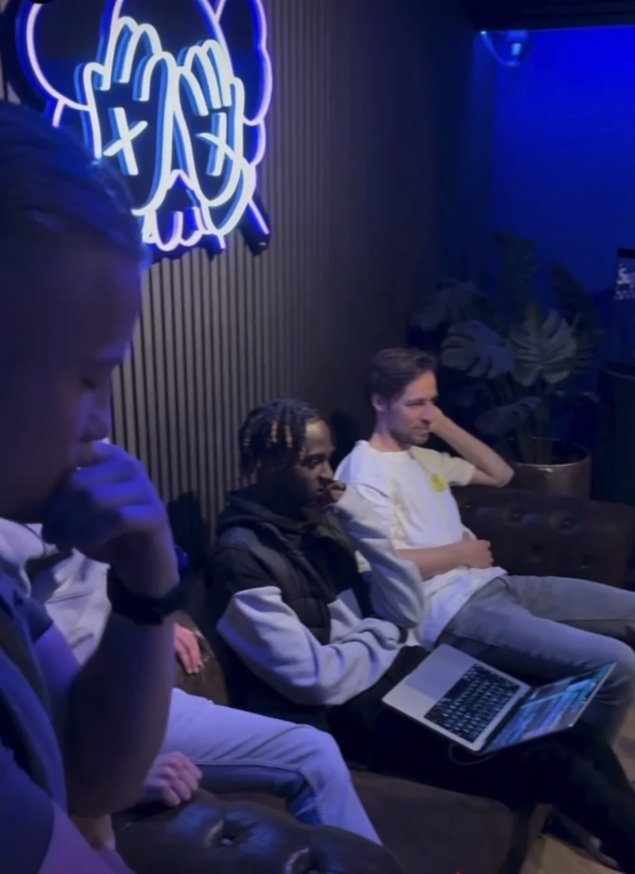 Three young men sitting in a dimly lit room with neon signage, one with a laptop, all appearing focused or thoughtful.