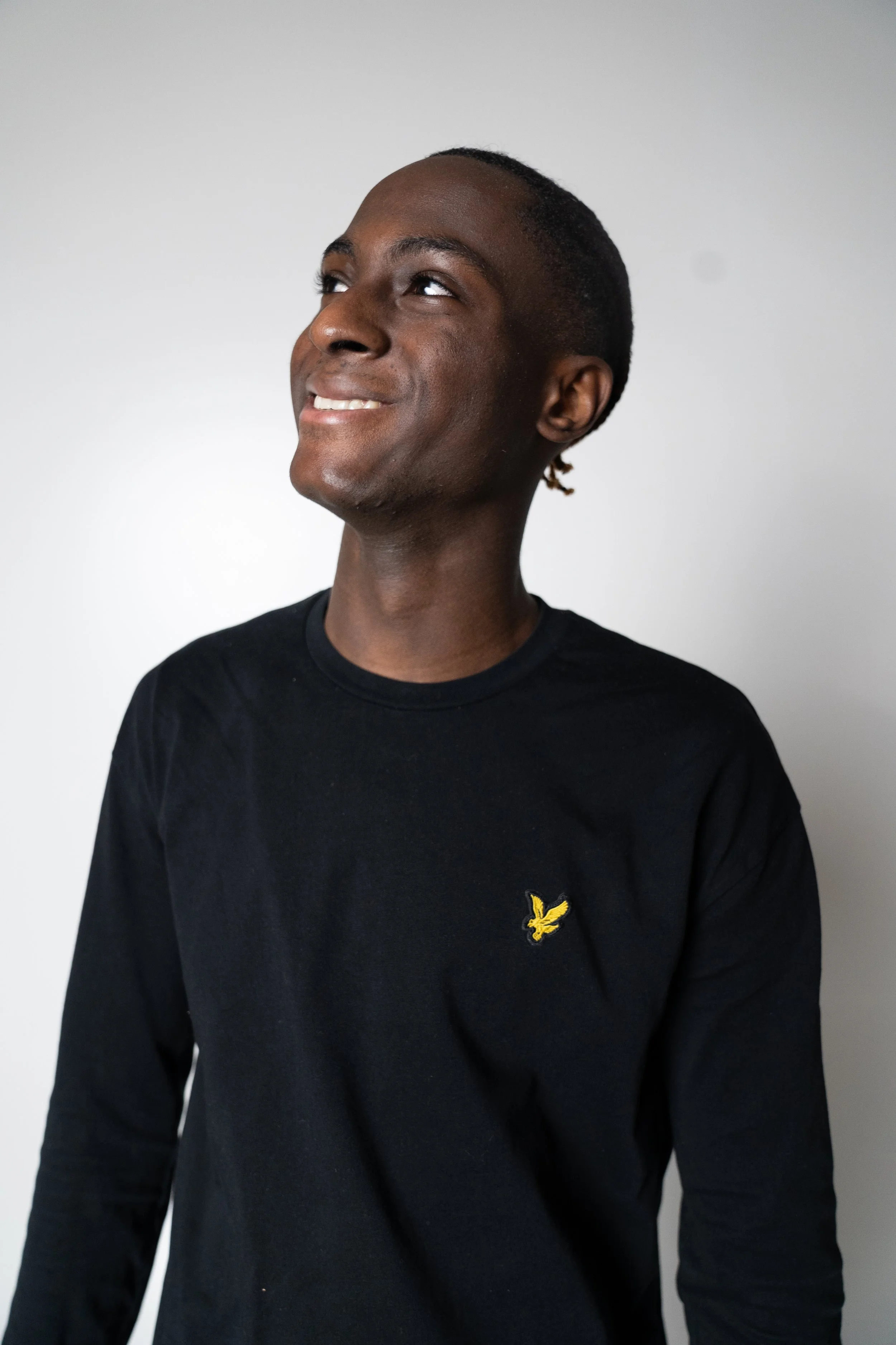 A smiling young man with short hair wearing a black long-sleeve shirt with a small yellow logo on the chest, standing against a plain white background.