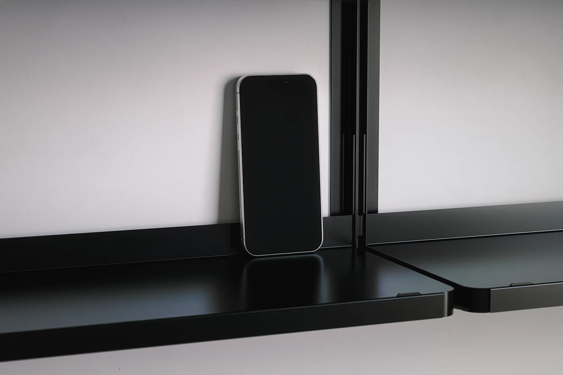 A smartphone resting on a black shelf against a white wall.