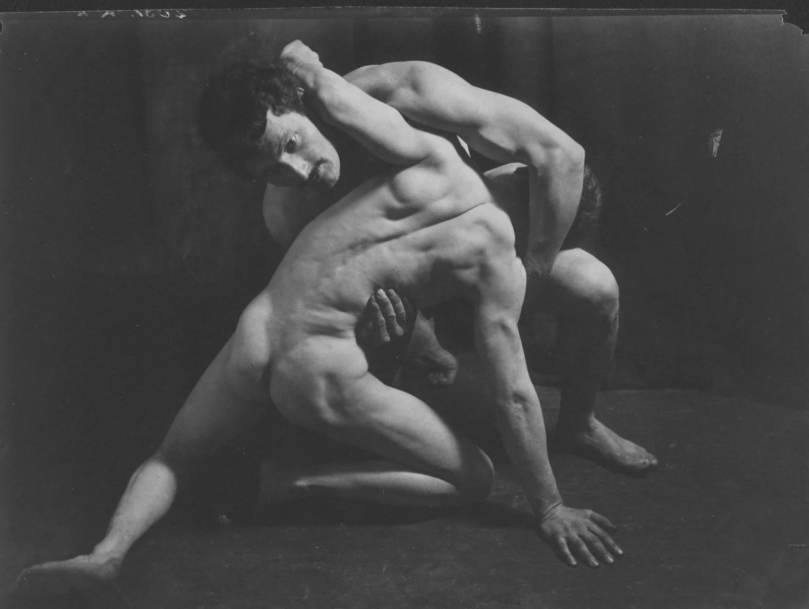 Grupo escultórico de dos luchadores, fotografiado en blanco y negro sobre fondo oscuro.