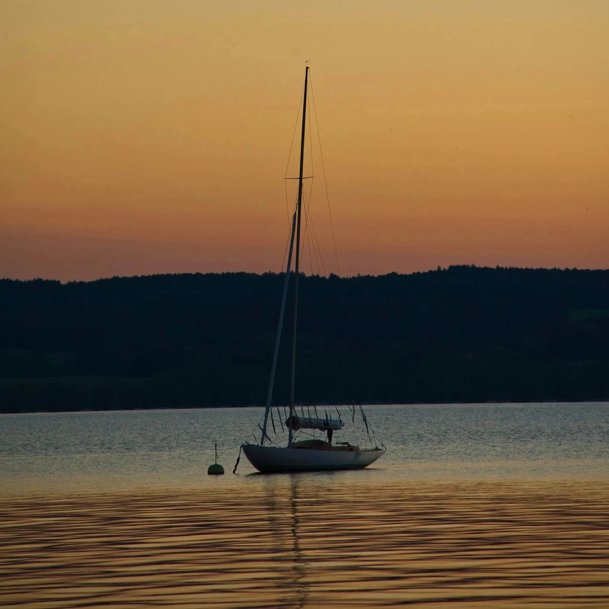 A sailboat anchored in calm water during sunset with a colorful sky and silhouette of distant hills.