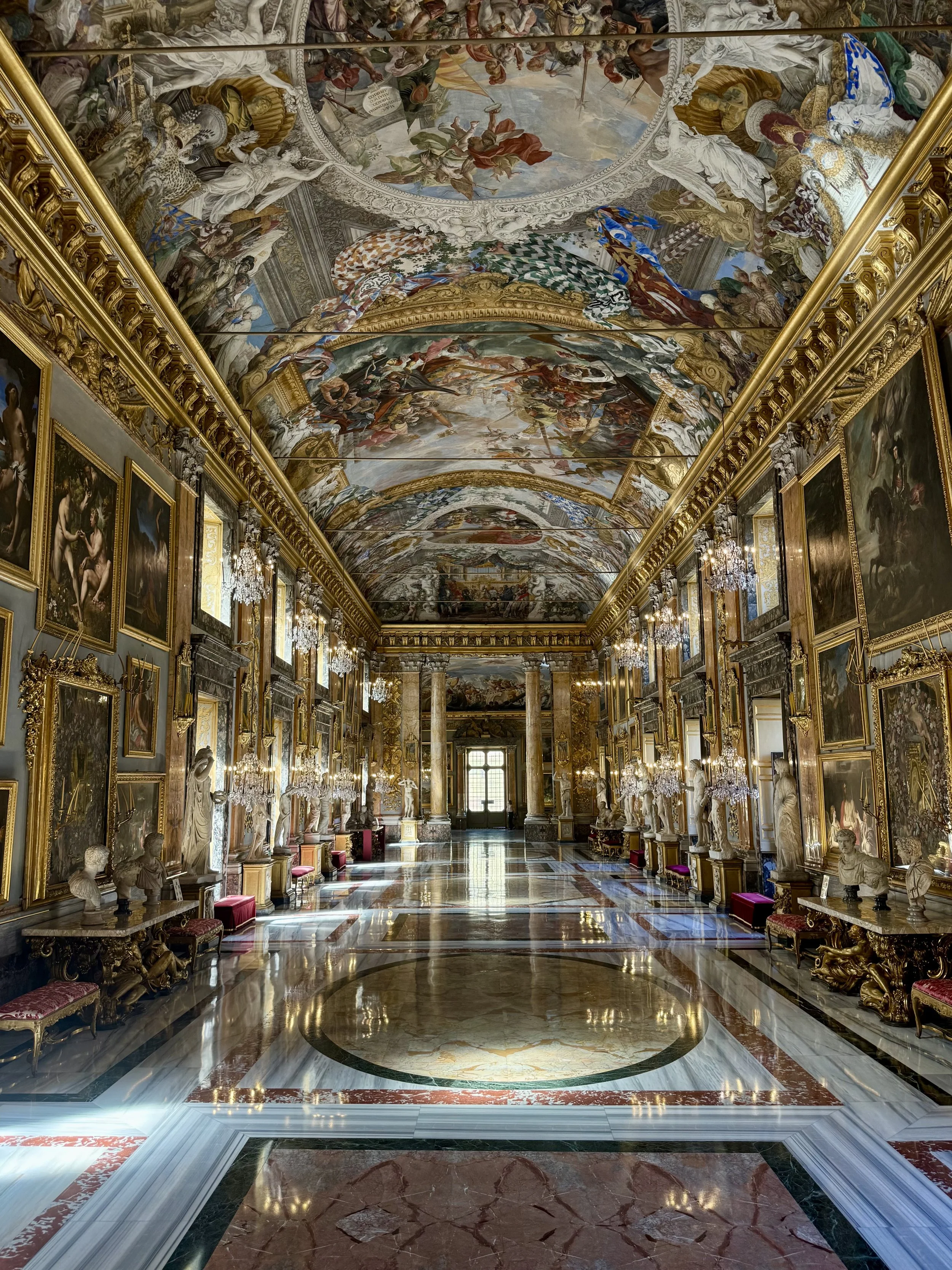 A grand, opulent gallery with ornate gold framing, marble bust sculptures, colorful paintings, chandelier lighting, and an intricately painted ceiling depicting mythological scenes.