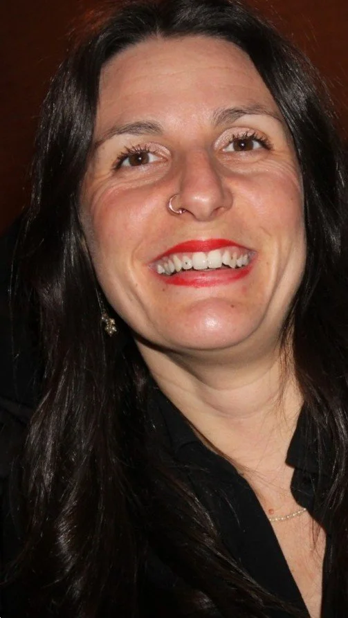 Portrait d'une femme avec les cheveux bruns, souriante, portant du rouge à lèvres rouge et une boucle d'oreille pendante, portant un haut noir.