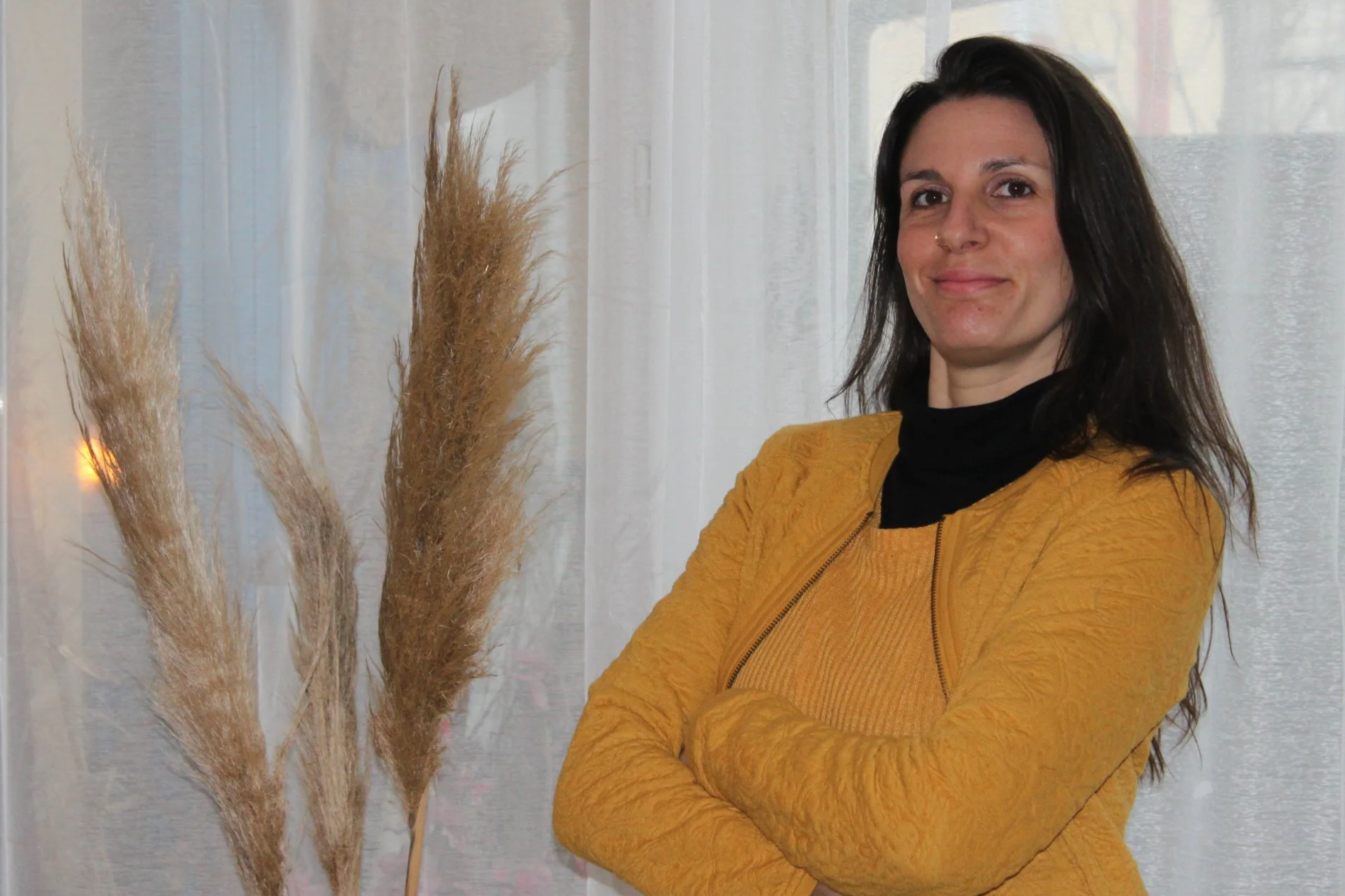 Une femme souriante avec des cheveux noirs, portant un pull noir et une veste jaune, pose avec ses bras croisés devant un décor intérieur avec des plumes naturelles et des rideaux blancs.