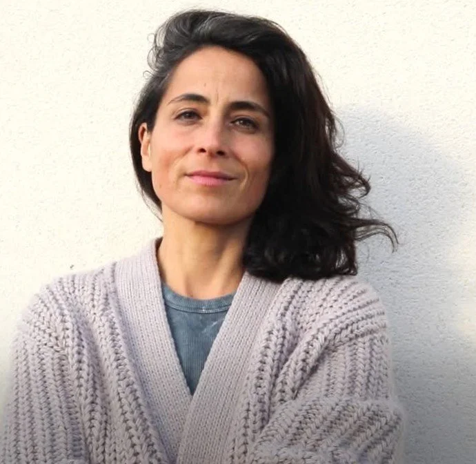Une femme avec des cheveux noirs et ondulés porte un pull gris en tricots et un t-shirt bleu, posant devant un mur blanc. Elle regarde l'appareil photo avec une expression neutre ou légèrement souriante.