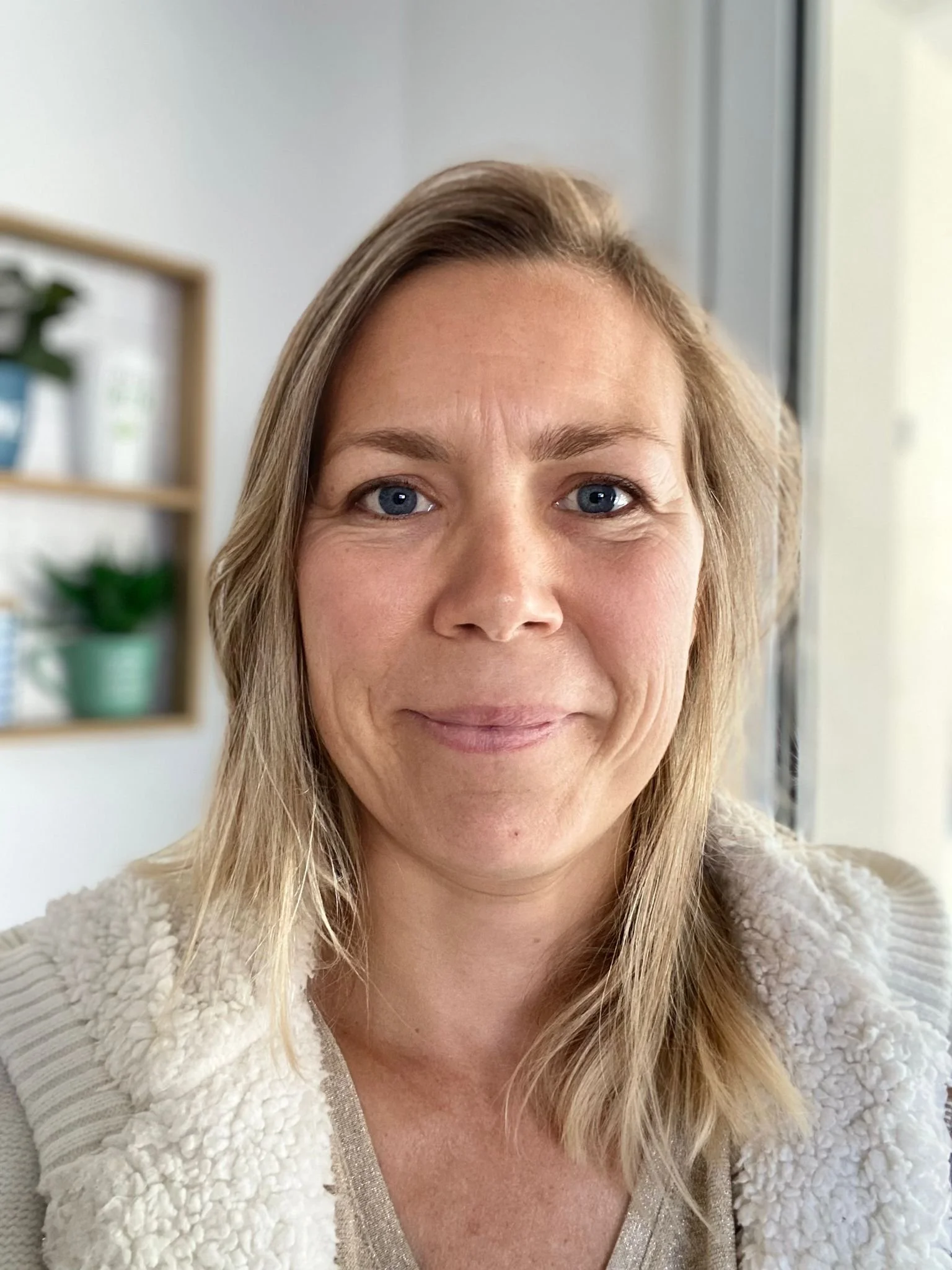 Une femme souriante à cheveux blonds, portant un pull beige, dans un intérieur lumineux avec des étagères contenant des plantes en arrière-plan.