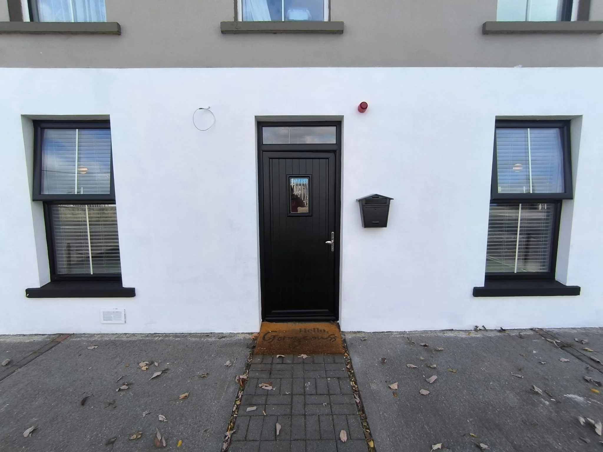 Front view of a white building with two large windows, a black door with a small window, a black mailbox, red alarm bell, and a welcome mat. Fallen leaves are on the sidewalk.