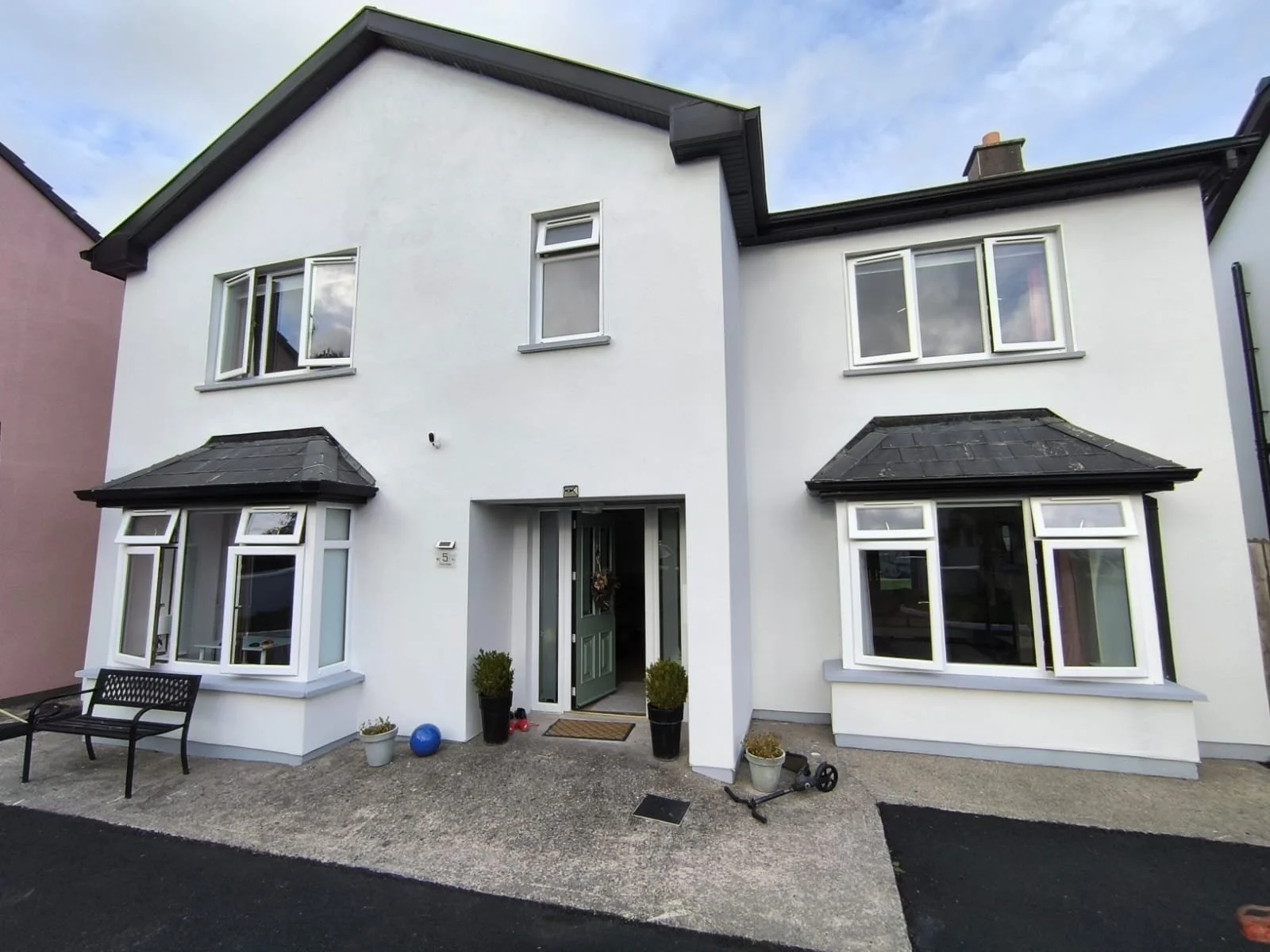 Front view of a white, two-story house with black roof, multiple windows, a green front door, and decorative plants outside.