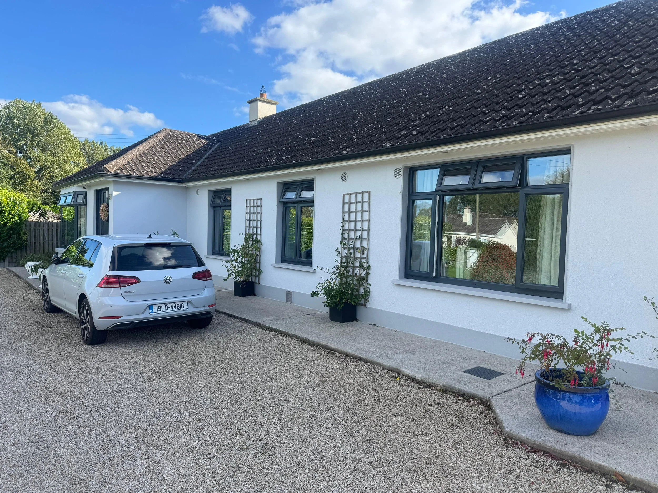 White house with black window frames, gravel driveway, Volkswagen Golf parked outside, flower pots with plants and flowers, blue flower pot, trees and sky in background.