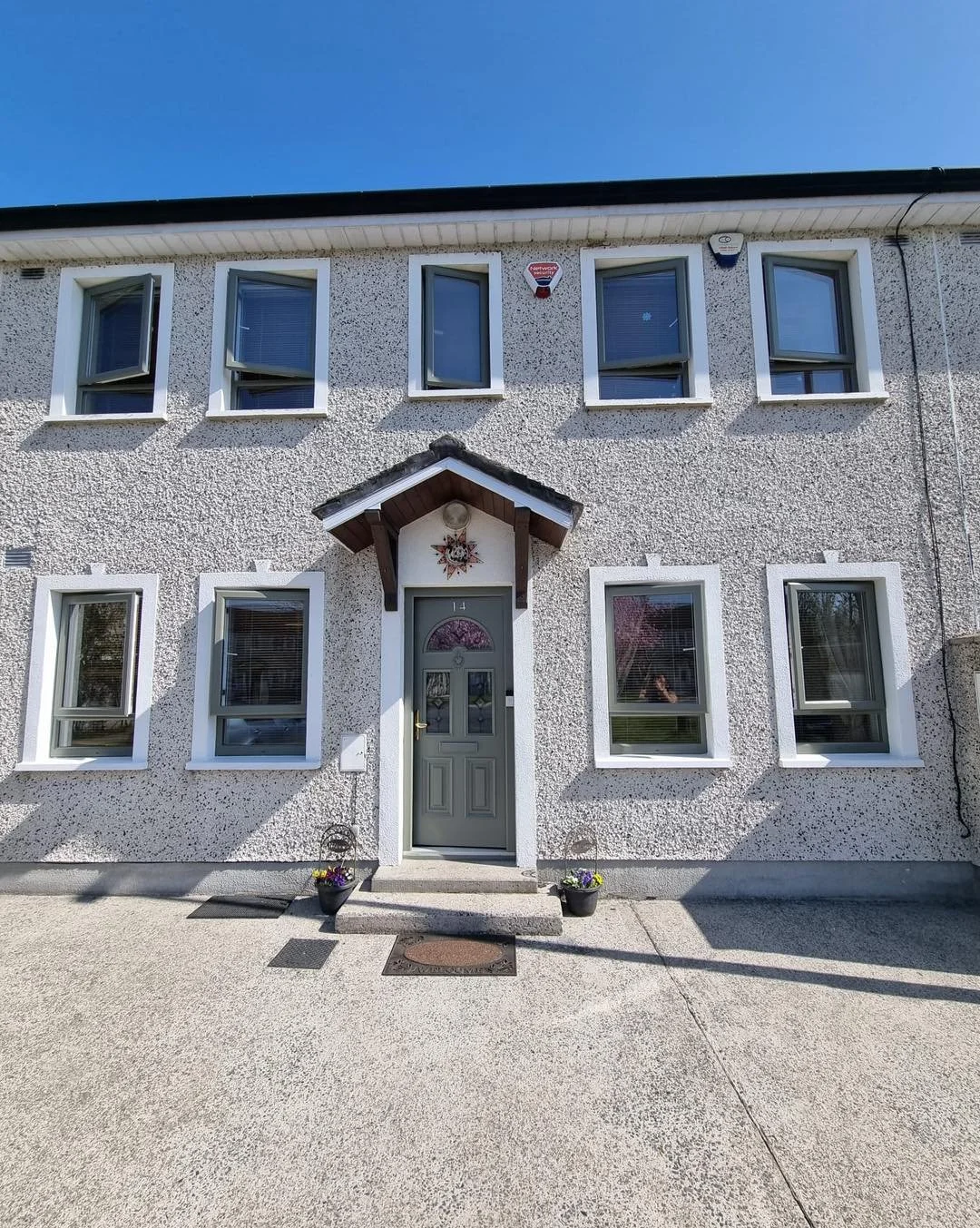 A two-story house with a textured white exterior, six windows with white frames, and a central front door with a small porch above it. The porch has a brown wooden overhang with decorative support brackets, and a star-shaped ornament adorns the front