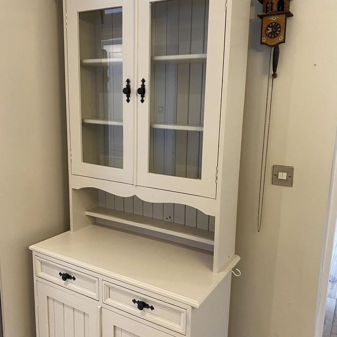 White wooden display cabinet with glass-paneled doors, black handles, and a lower section with drawers, situated against a beige wall with a wall clock and a light switch nearby.