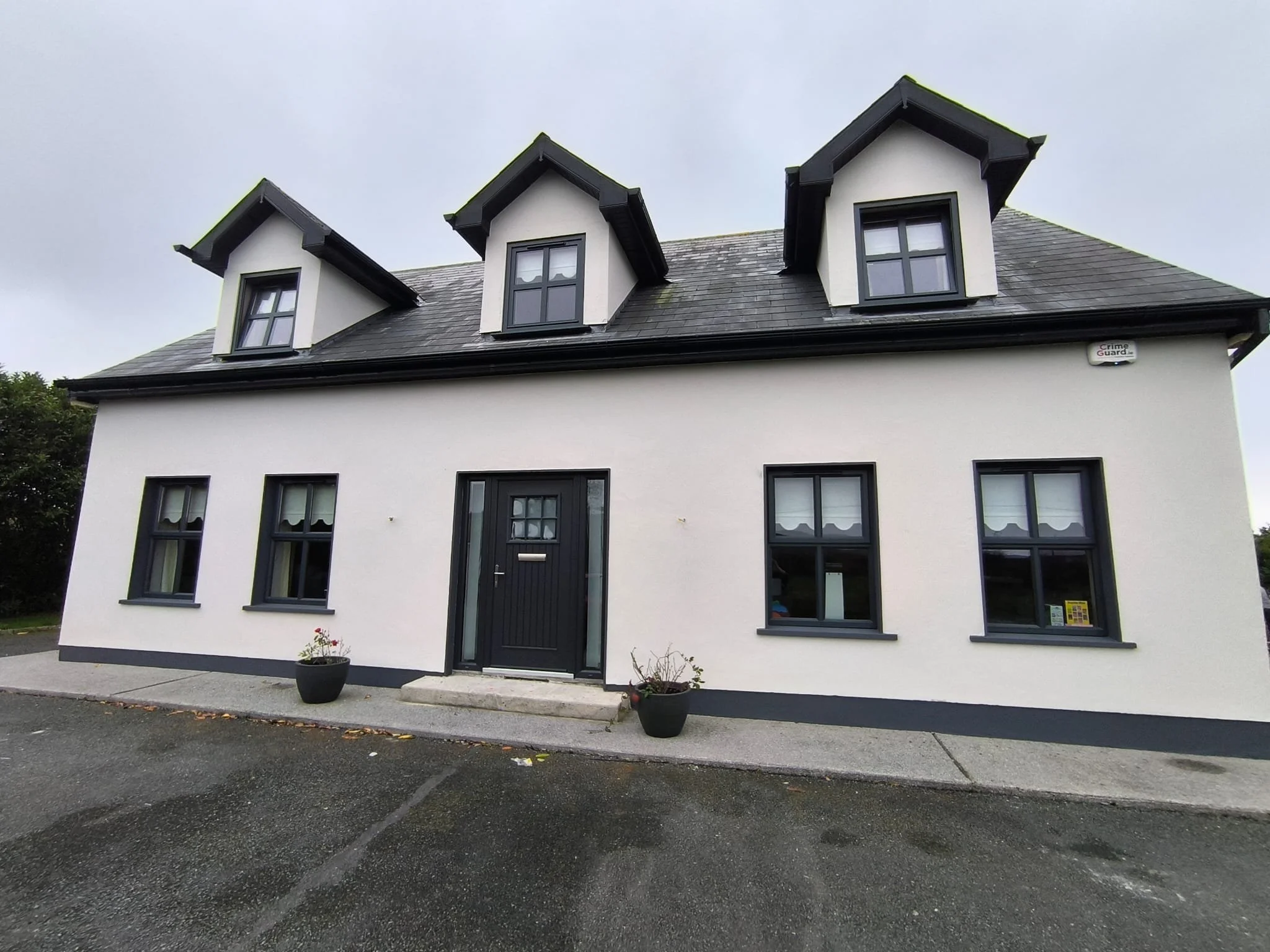 Modern white house with black-trimmed windows, black front door, and three dormer windows on the roof, with a parking area in front and trees in the background.