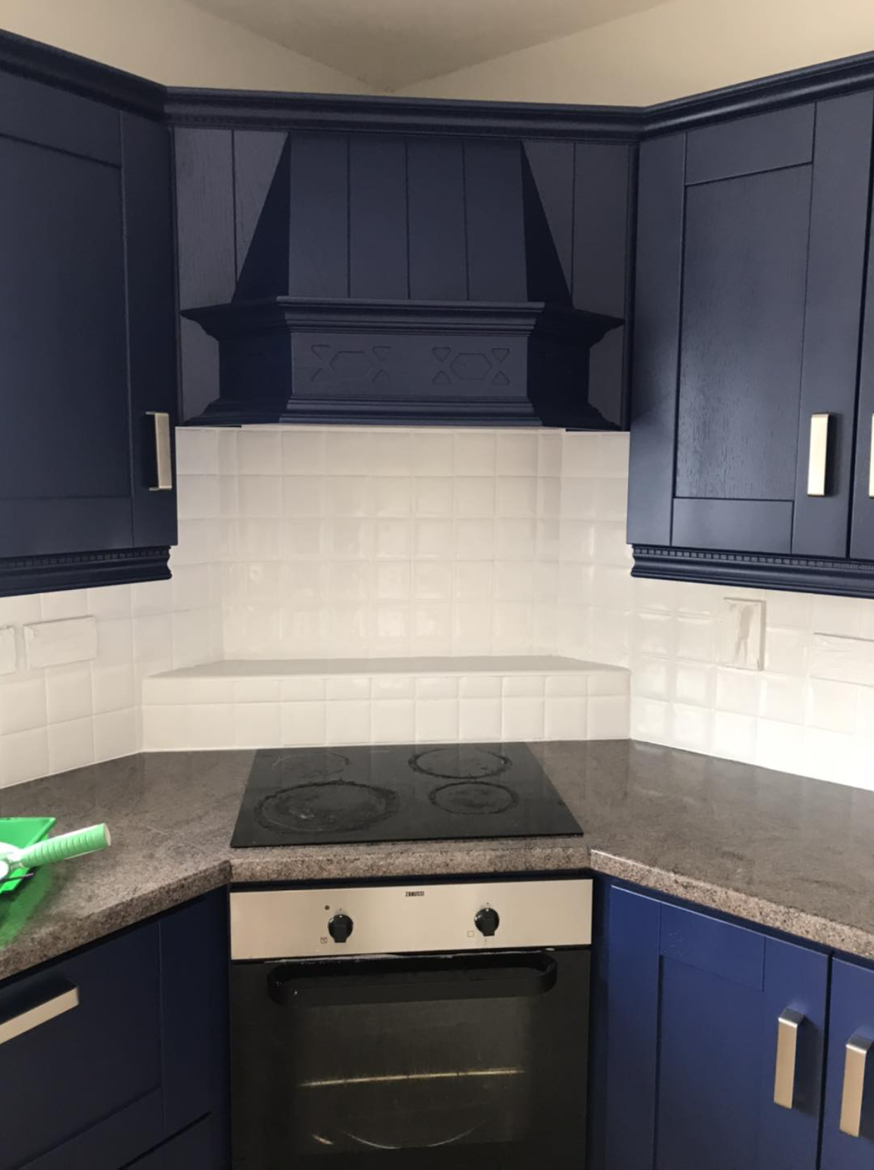 Corner of a kitchen with dark blue cabinets, a black cooktop, white tiled backsplash, and a black range hood.